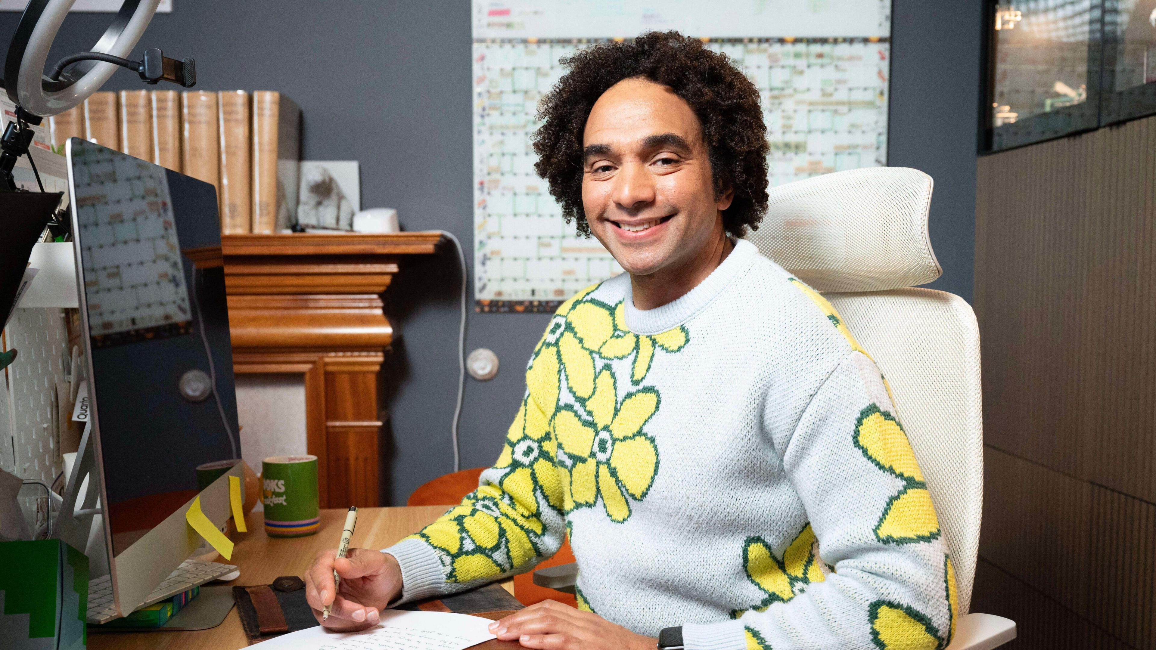 Joseph Coelho has curly hair and is wearing a grey and yellow jumper. He is sat at a desk.