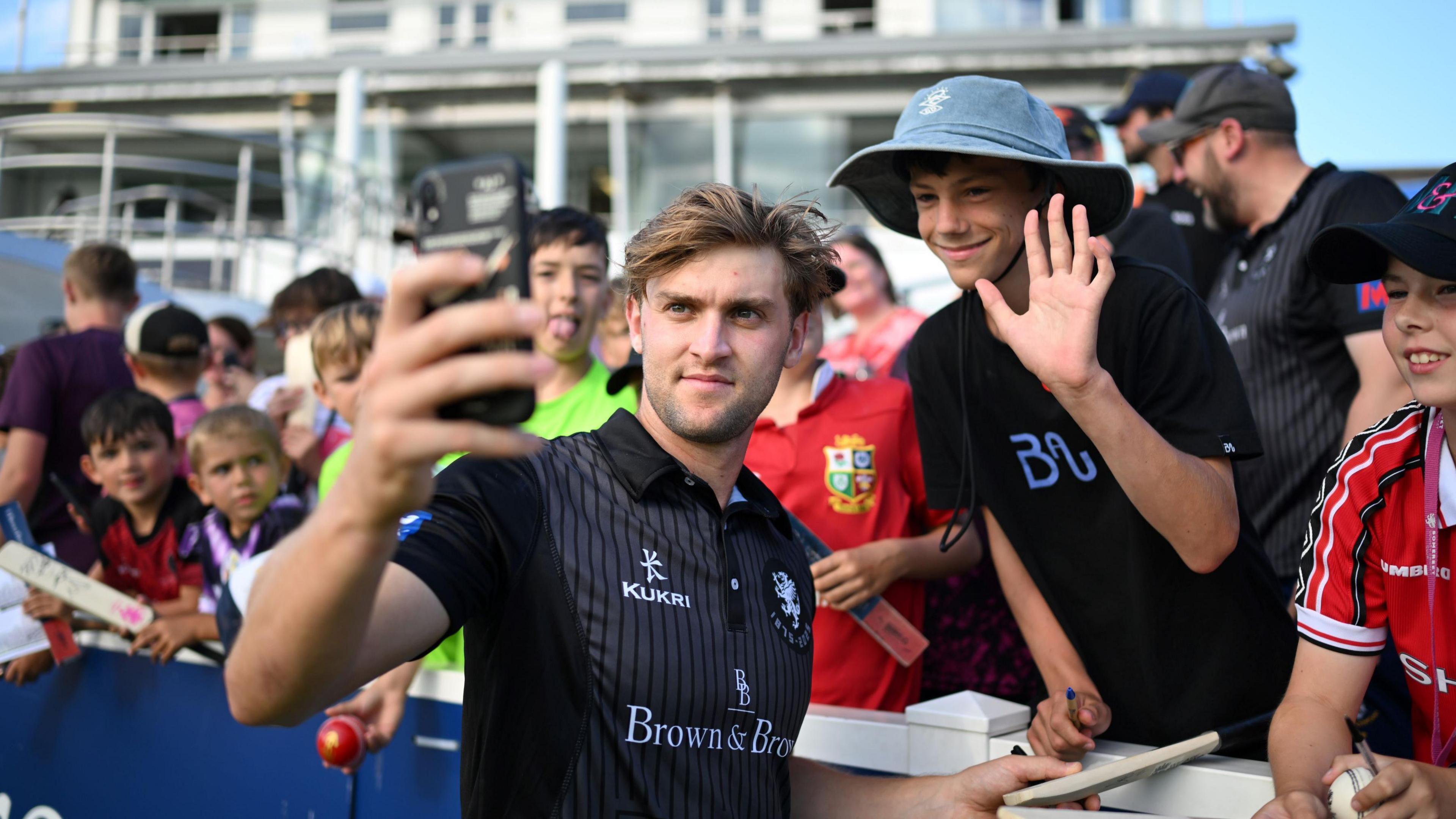 A fair-haired man standing in front of a group of boys. The man at the front is wearing a black cricket polo shirt which has three logos on its front. He is holding a phone up to take a selfie. There is a young male fan wearing a hat standing next to him for the photo. He has his hand up in a wave and is smiling.