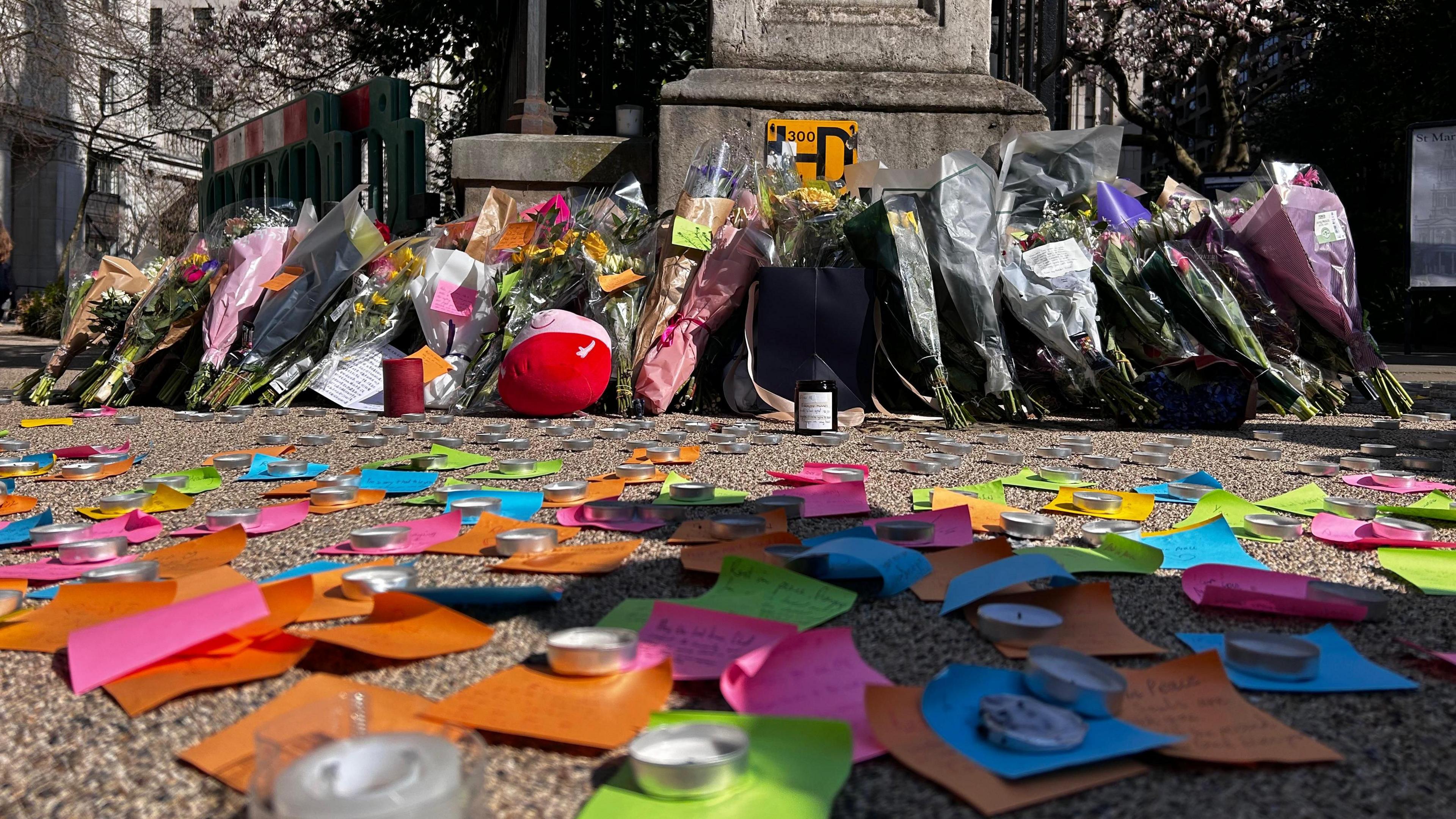 Multi-coloured sticky notes on the pavement, with tributes written on them, and tea lights on top, leading up to a row of flowers.