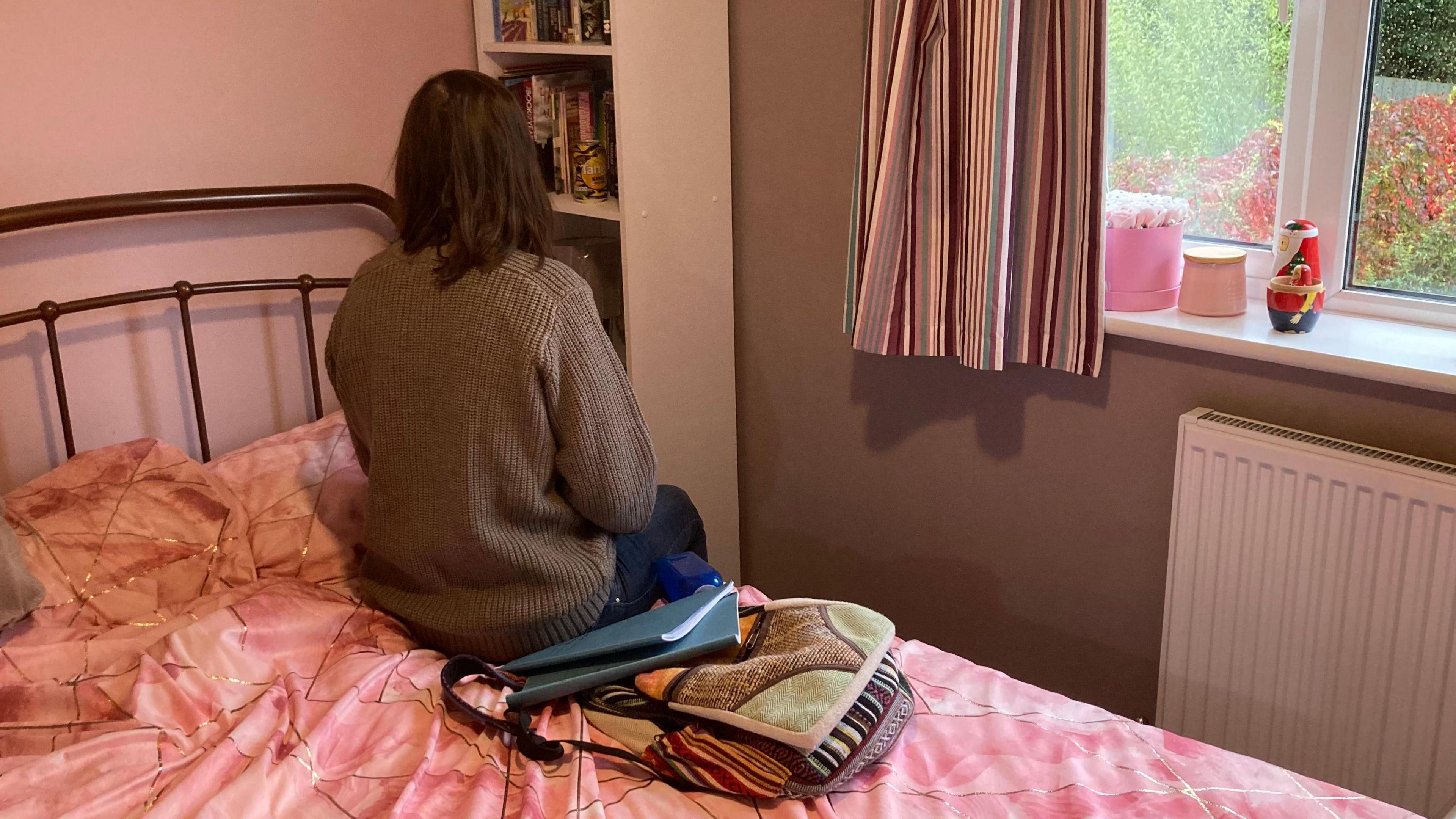 The image shows the back of a young person with long brown hair. They are wearing a brown sweater and are sitting on a bed with a pink duvet. There is a rucksack with exercise books on the bed. There is a bookshelf to their right and striped curtains to the left of a window.