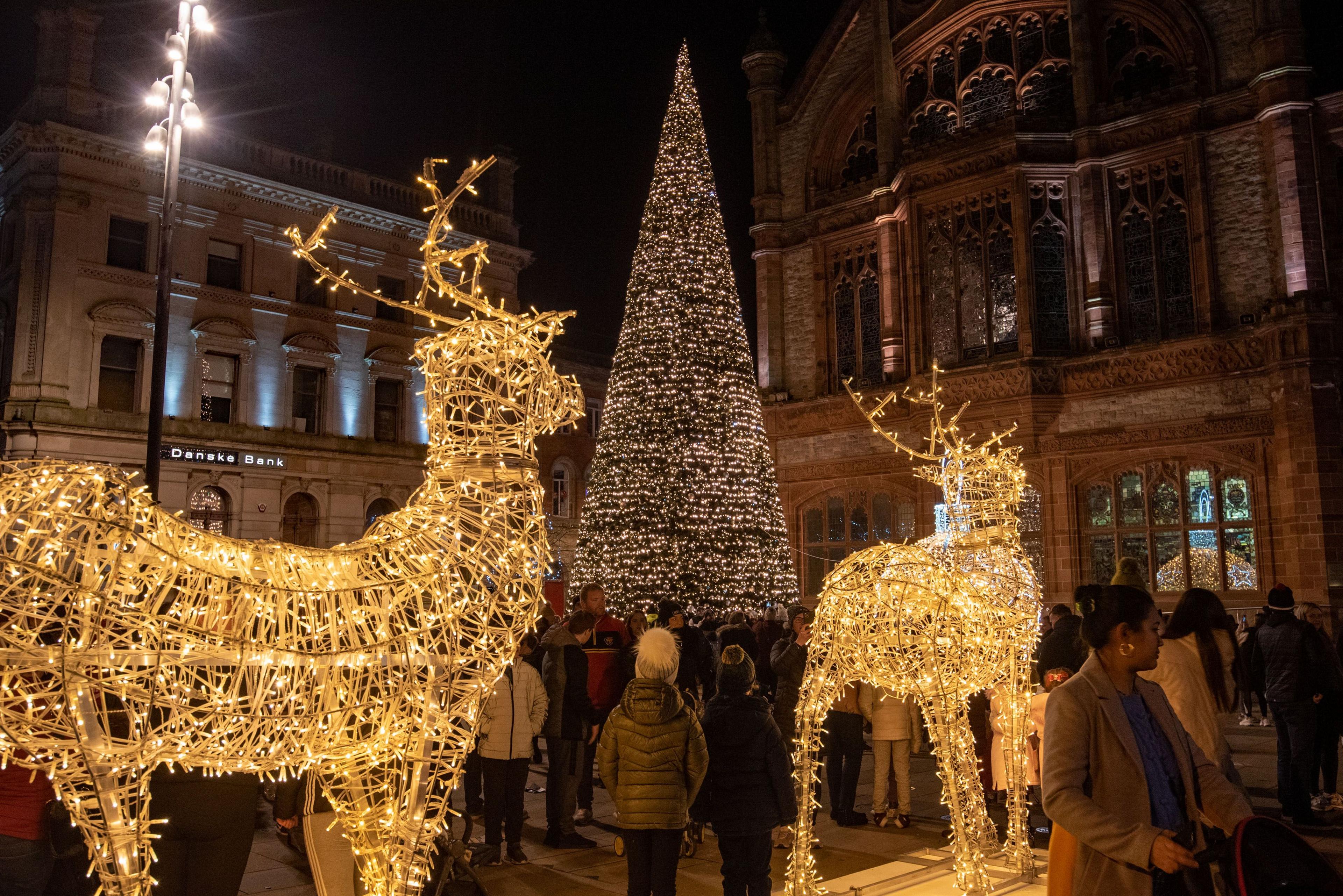 Two illuminated reindeer decoration are positioned behind a civic Christmas tree in Derry which is also lit up. The Guildhall is in the backgound. A number of people are gathered around the decorations.