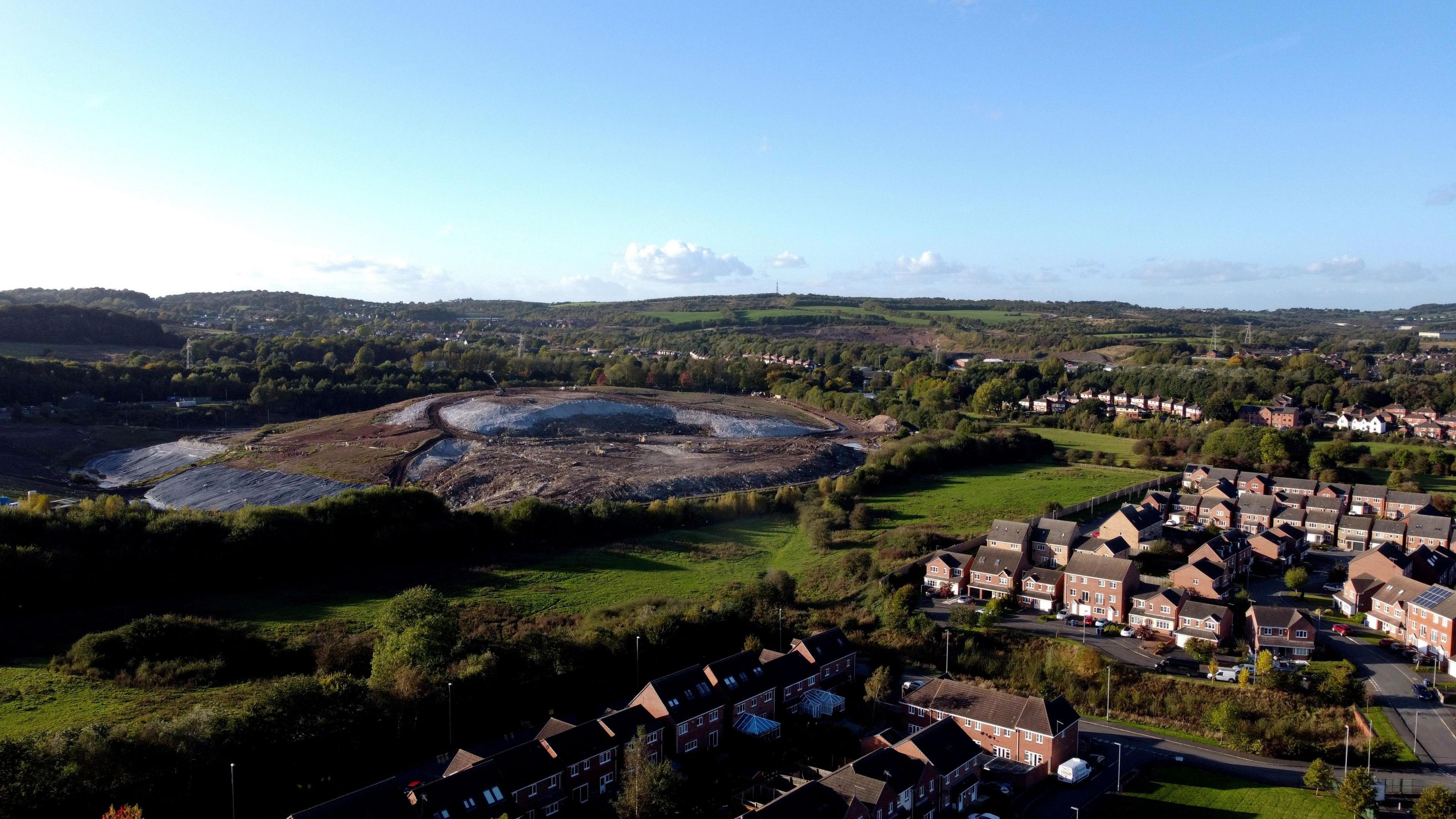 An aerial drone view of a town with houses on the right, and a large landfill site on the left