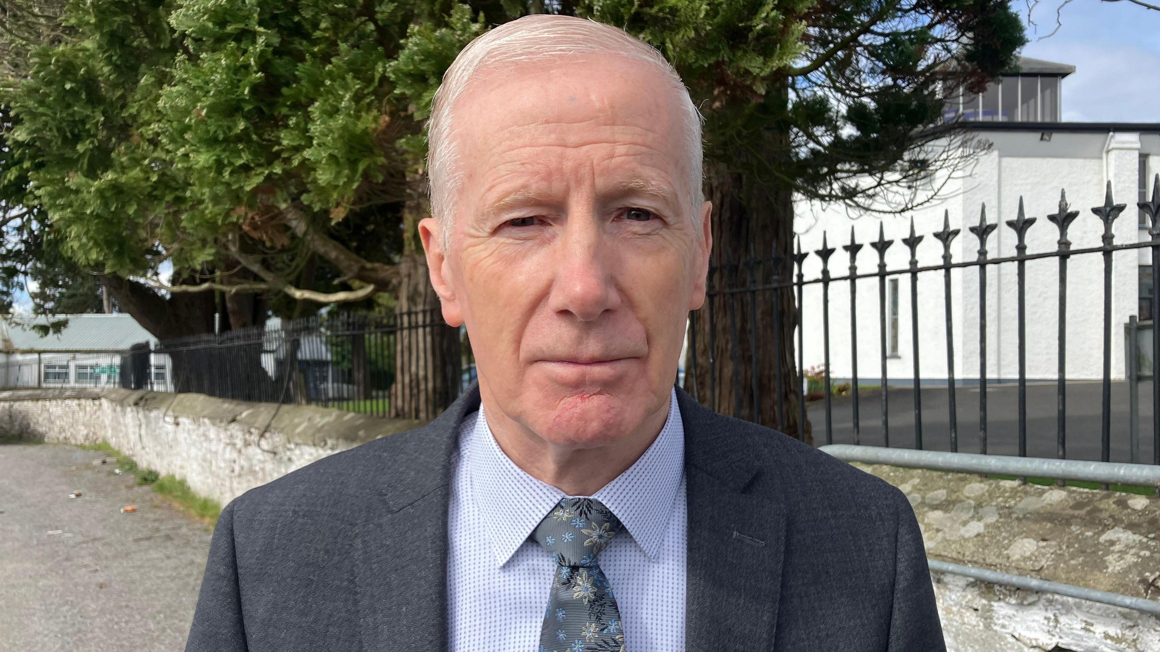 A man stands close to the camera, with short white hair, wearing a grey suit and blue shirt.