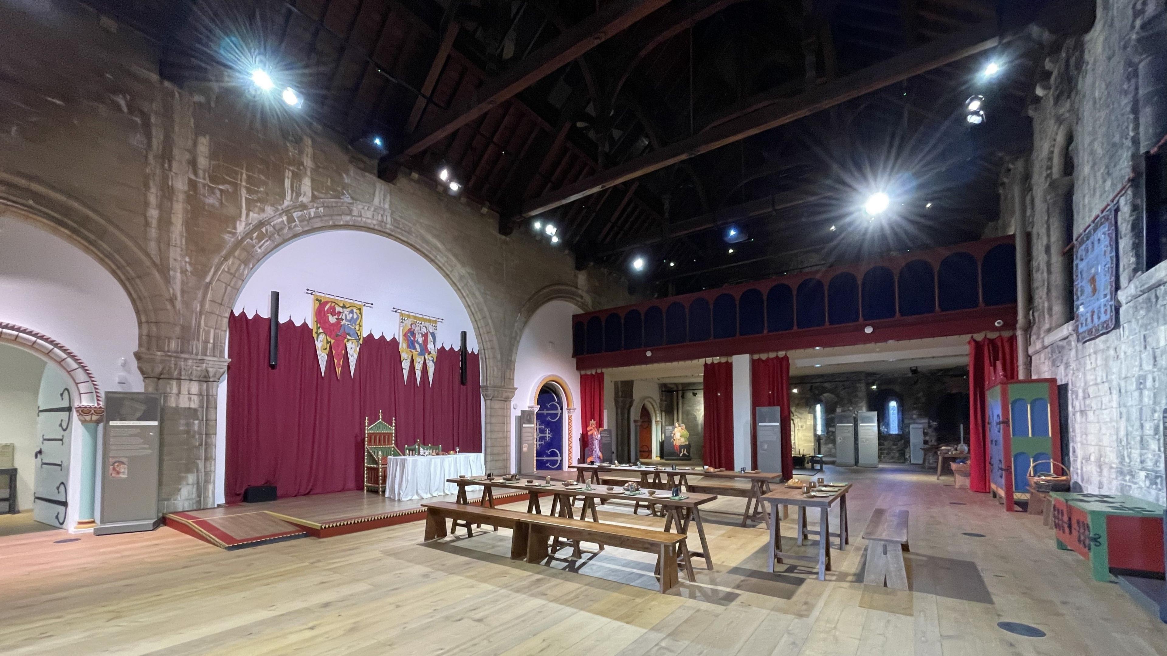 A recreation of how a Norman castle's great hall would have looked, with a coat of arms hanging above the king's table and with other tables laid out for dining in front of them, with high wooden ceilings above.