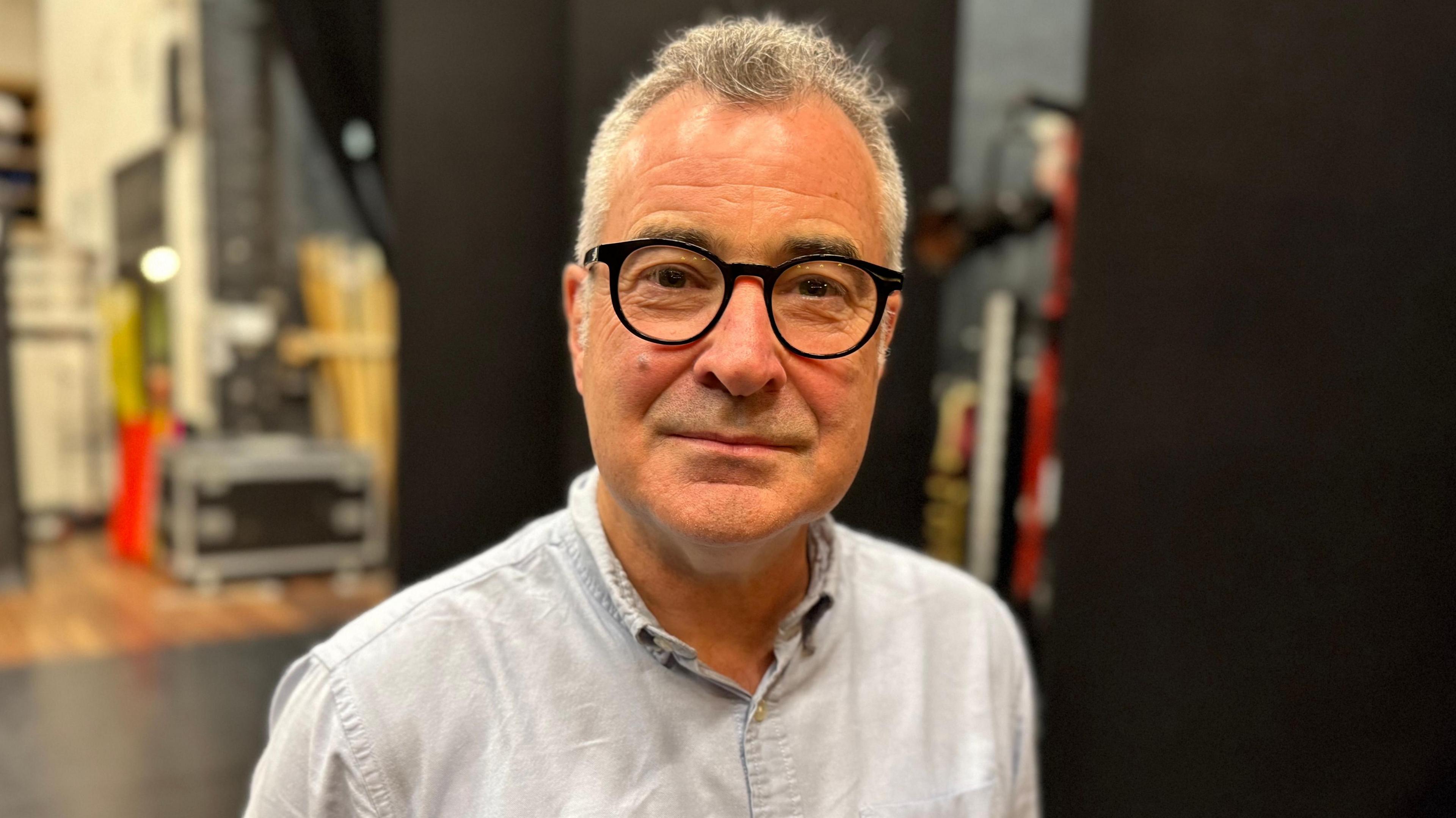 Séamus Shea wears a blue shirt and black-rimmed glasses. He has grey hair and stands in front of two black backdrops backstage. You can also see boxes and props blurred out in the background.