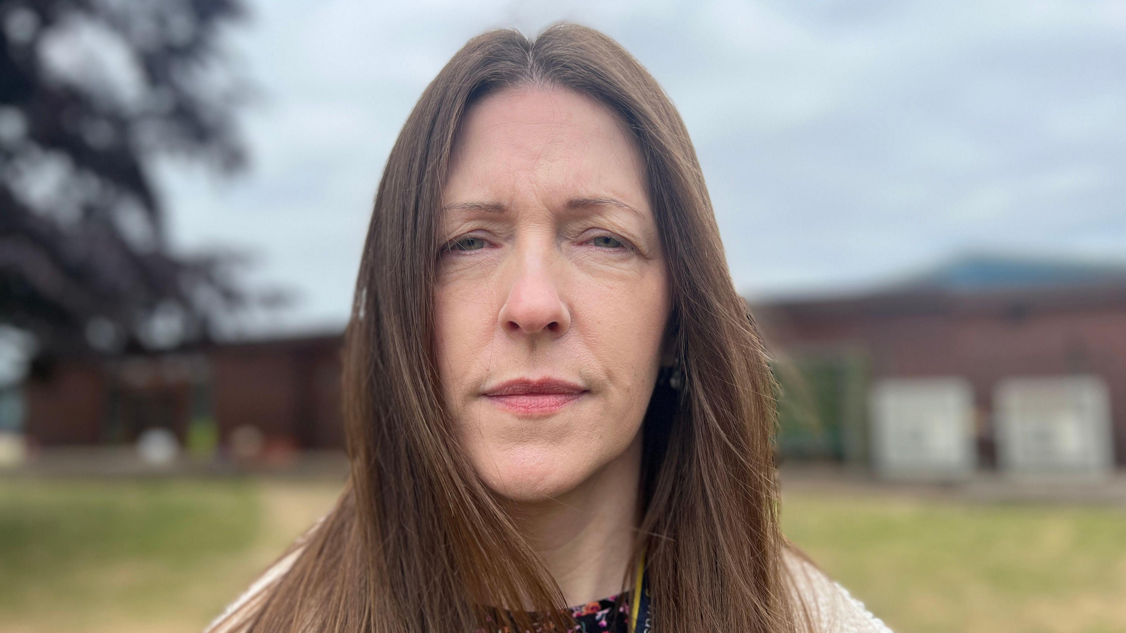 Fran Rhodes has long brown hair and is wearing a white fluffy coat. The school field behind her has been blurred.