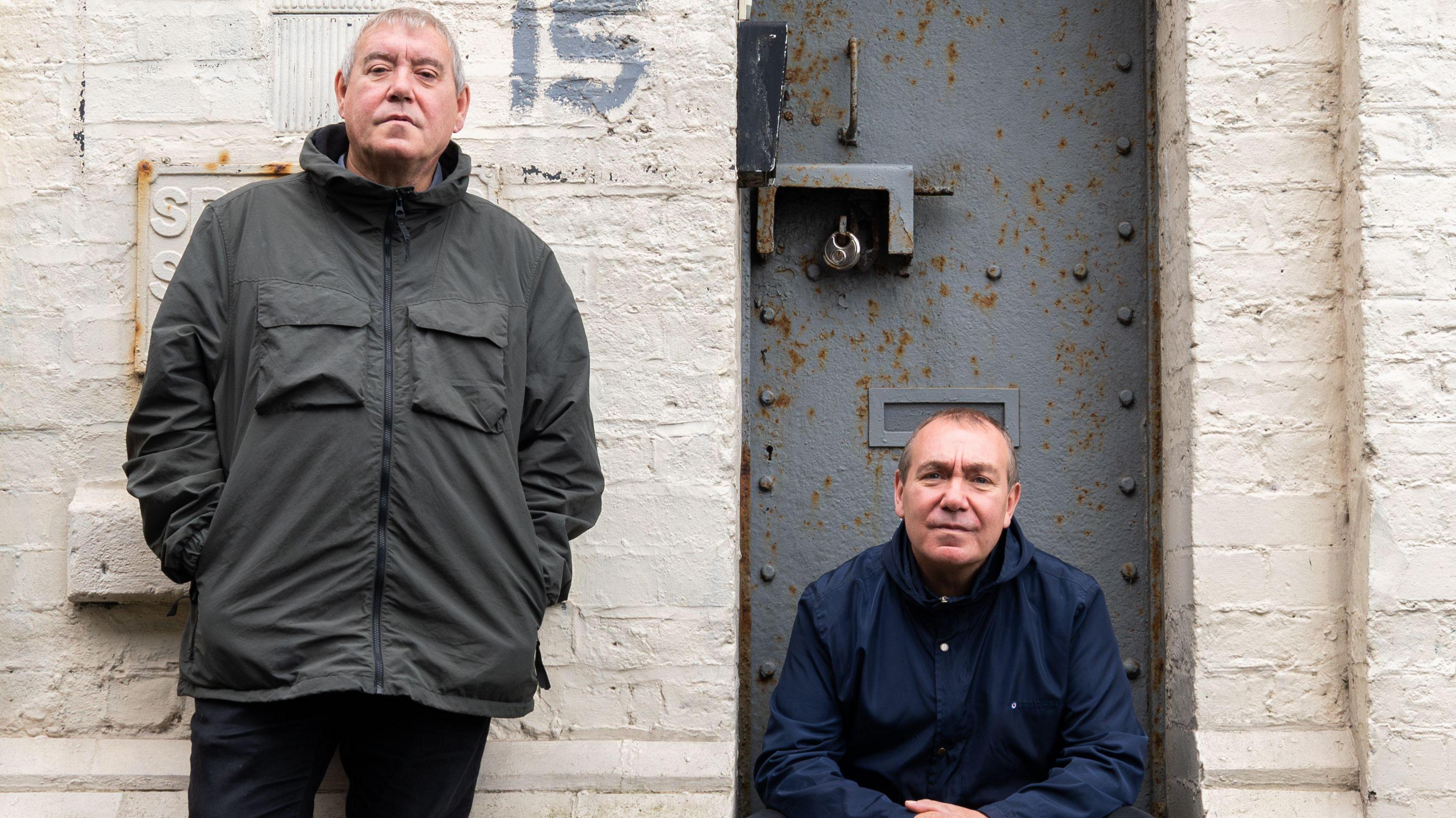 Tony and Chris Griffiths outside their studio in Birkenhead. Tony leans against a white brick wall while Chris sits on the door step. 