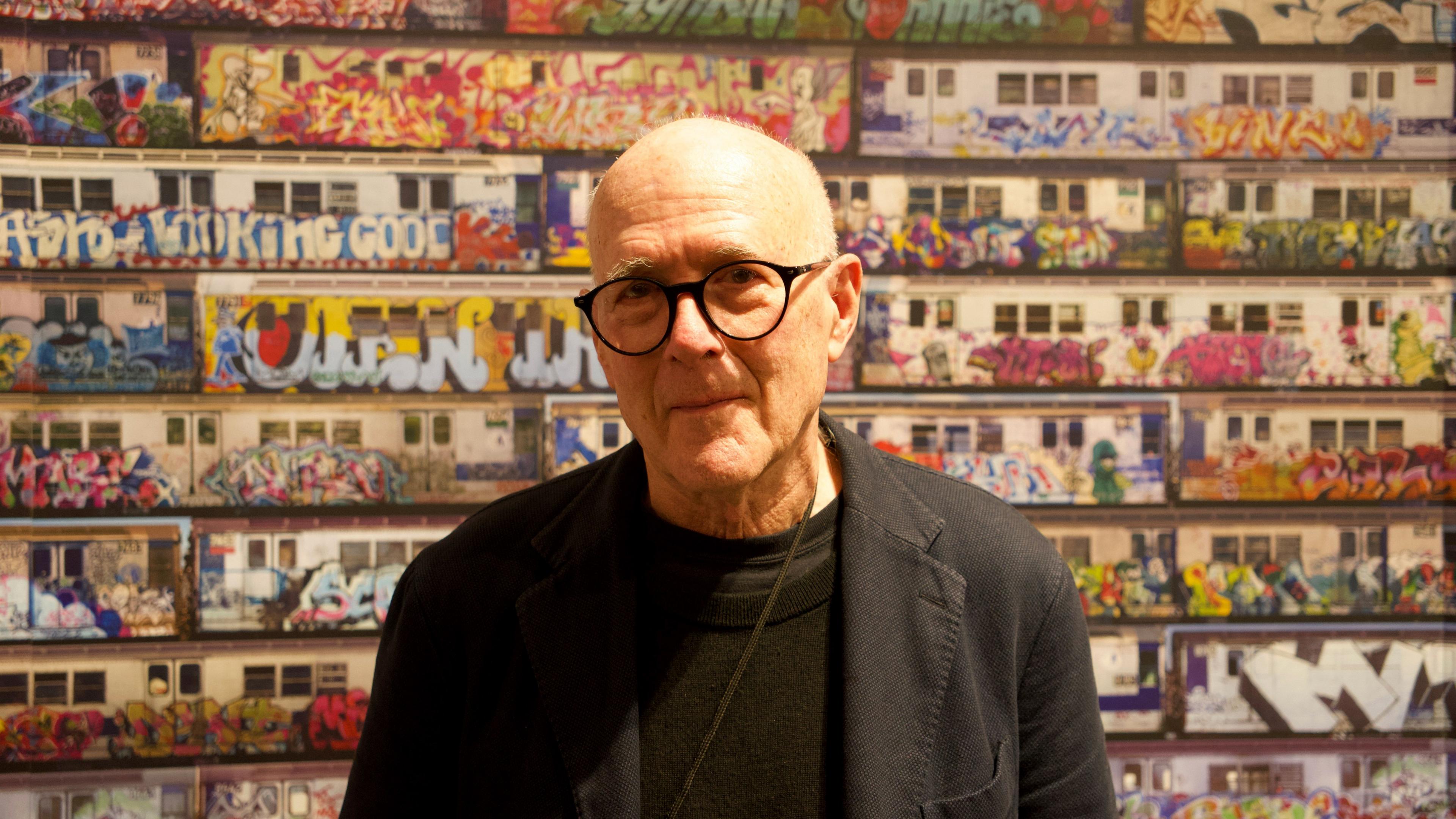 A balding man with glasses and dark jumper and jacket stands in front of a wall with subway train images across it.