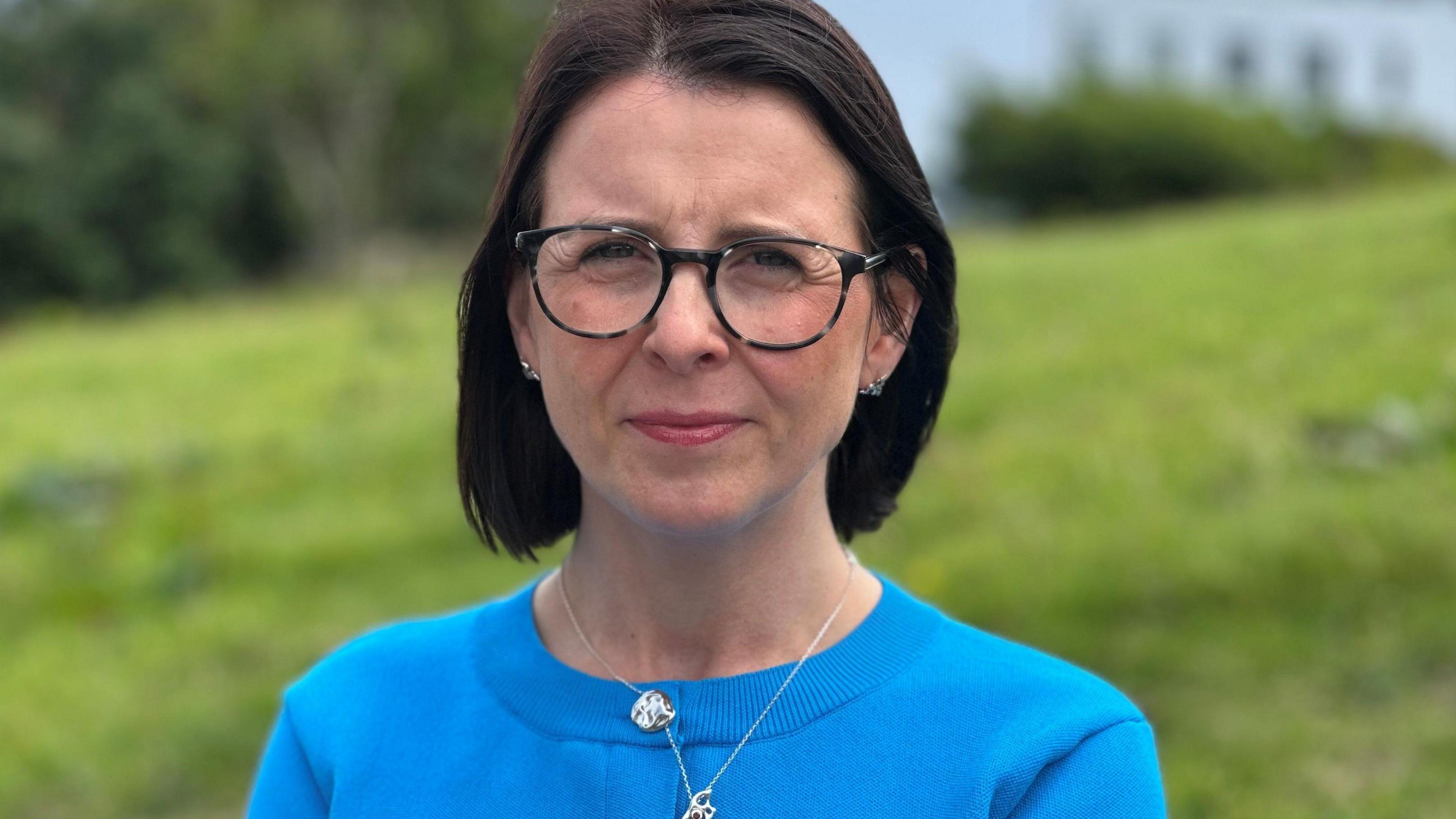 Sarah Maltby has short dark hair and wears a blue cardigan and silver necklace. She also wears dark glasses. She stands in front of a grassy bank.