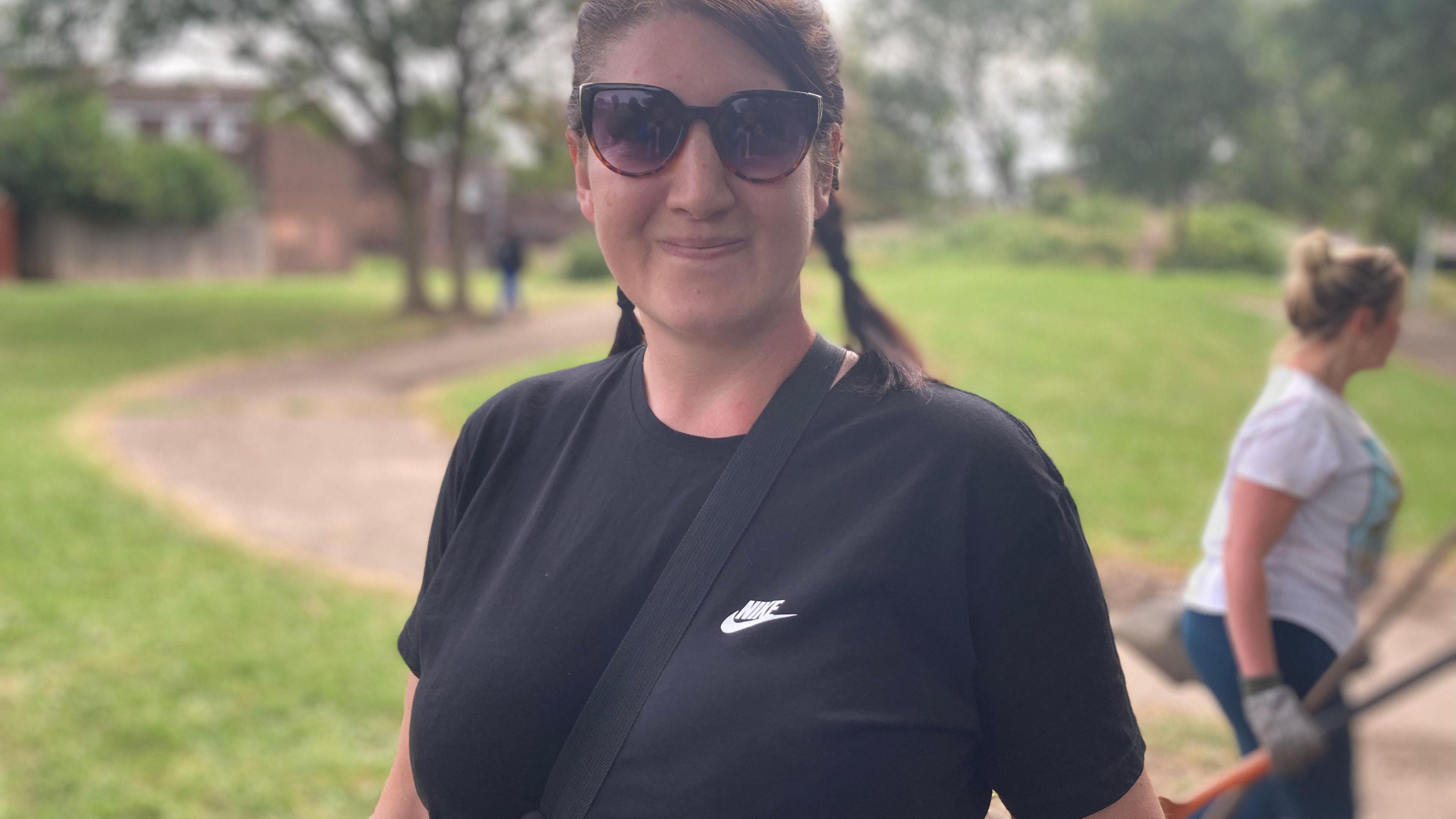 A woman wearing sunglasses and a black Nike T-shirt in a park with grass and trees in the background.