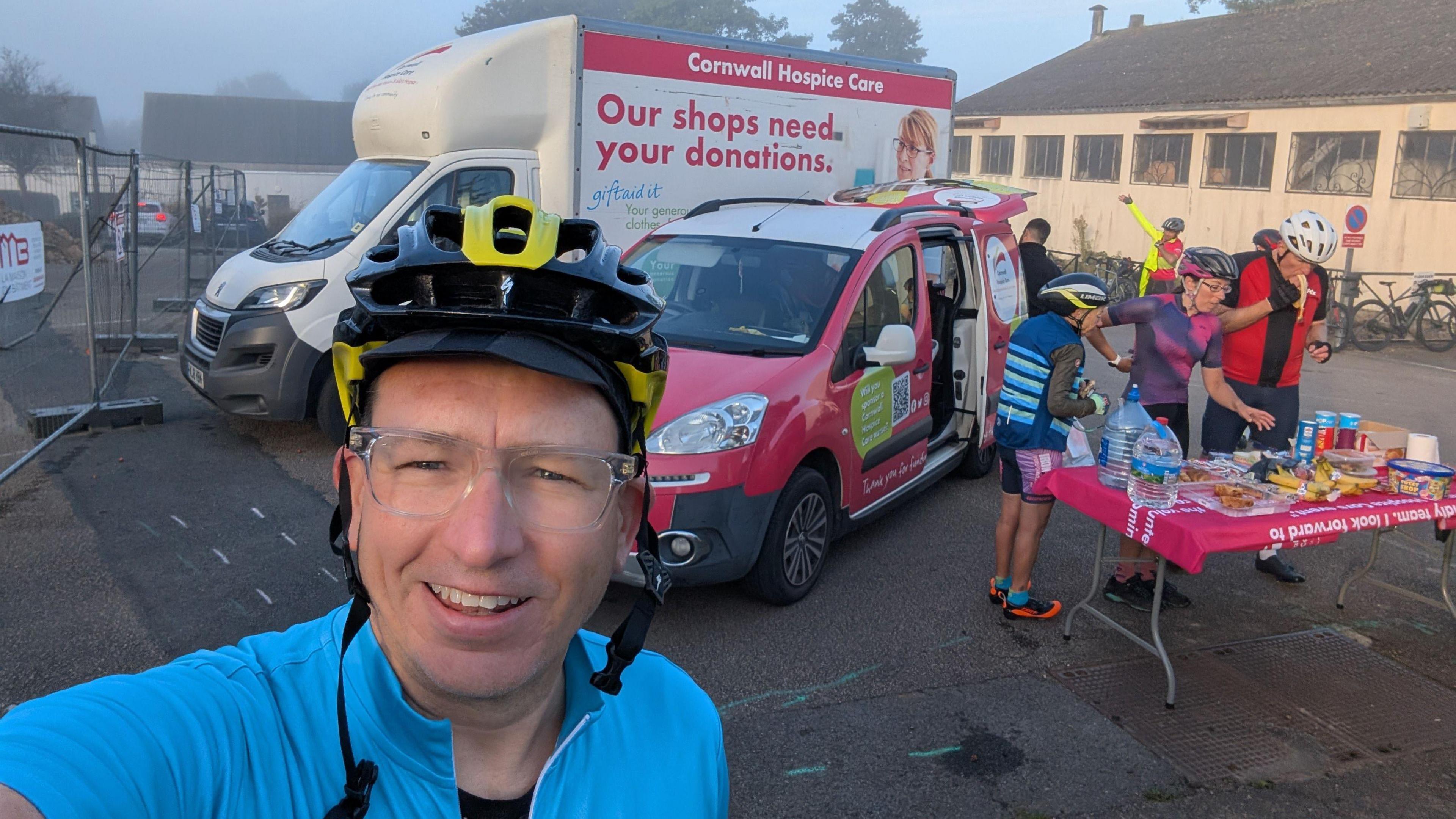 A man taking a selfie wearing cycling gear with Cornwall Hospice Care vans behind him and people at a table eating snacks. Foggy skies and buildings behind.