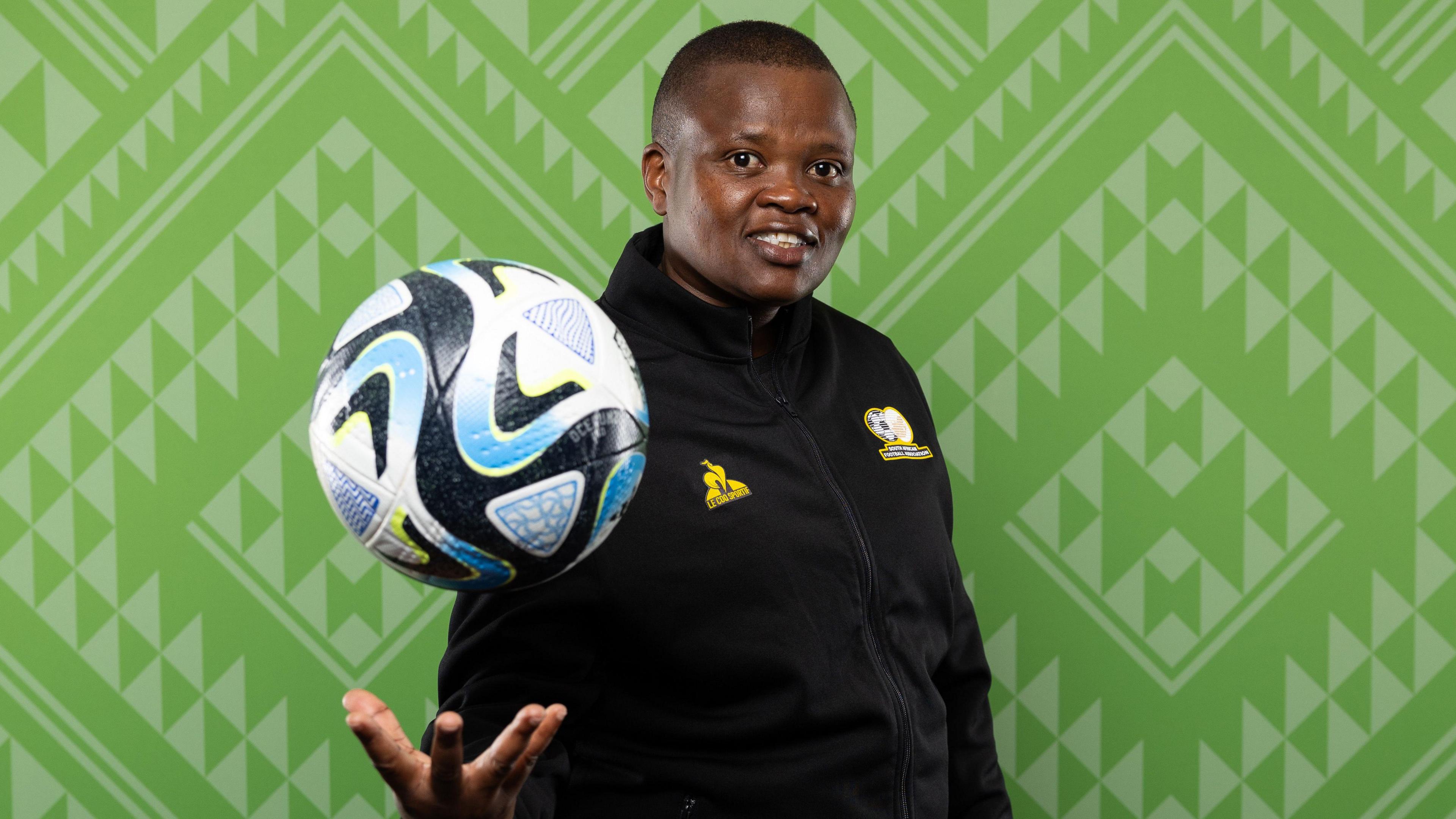 Thinasonke Mbuli, wearing a black South African training kit, throws a ball up into the air and smiles at the camera while standing in front of a wall bearing a green geometric pattern