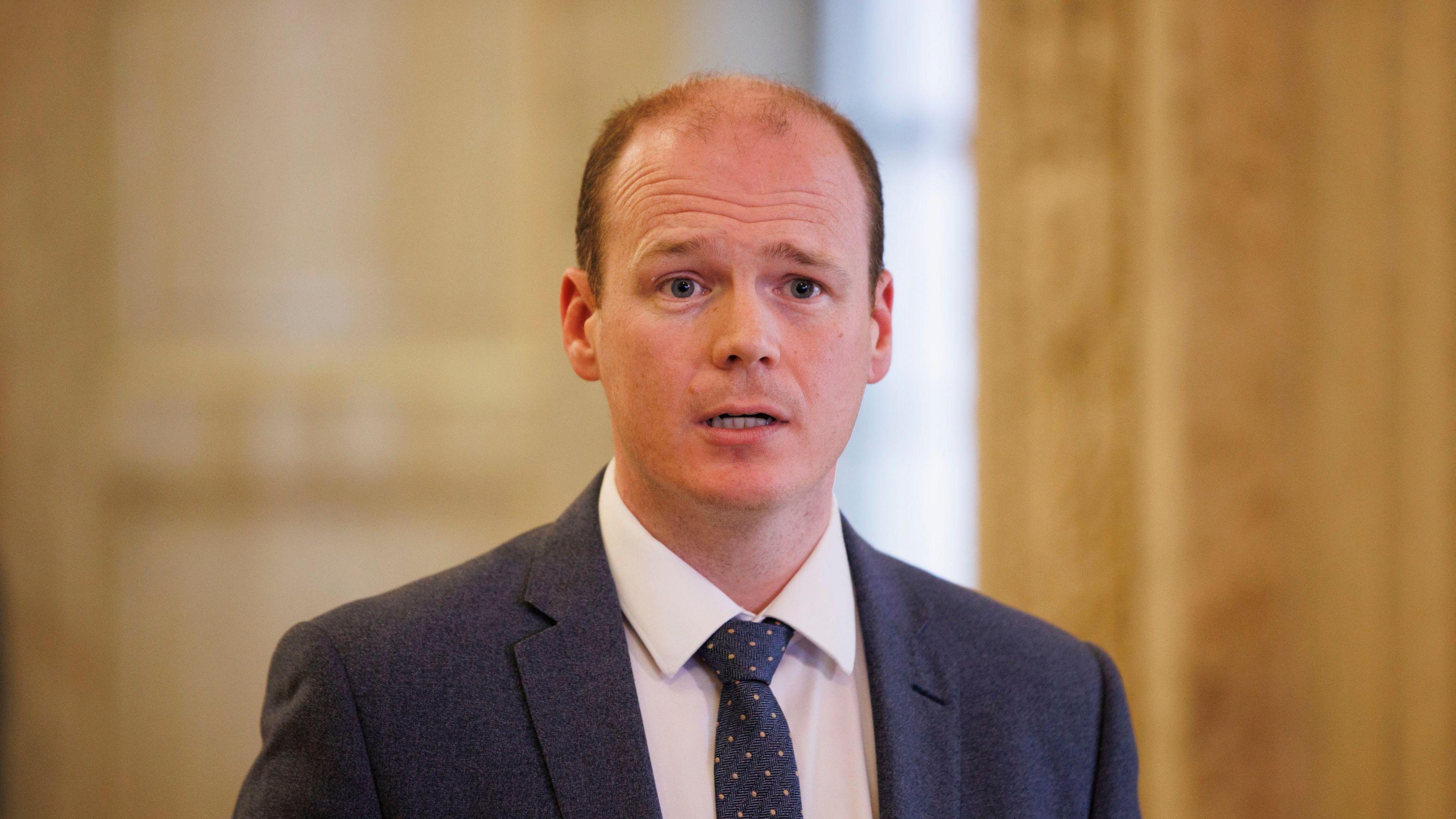 Gordon Lyons, with short brown hair, wearing a white shirt, navy tie and navy suit jacket.