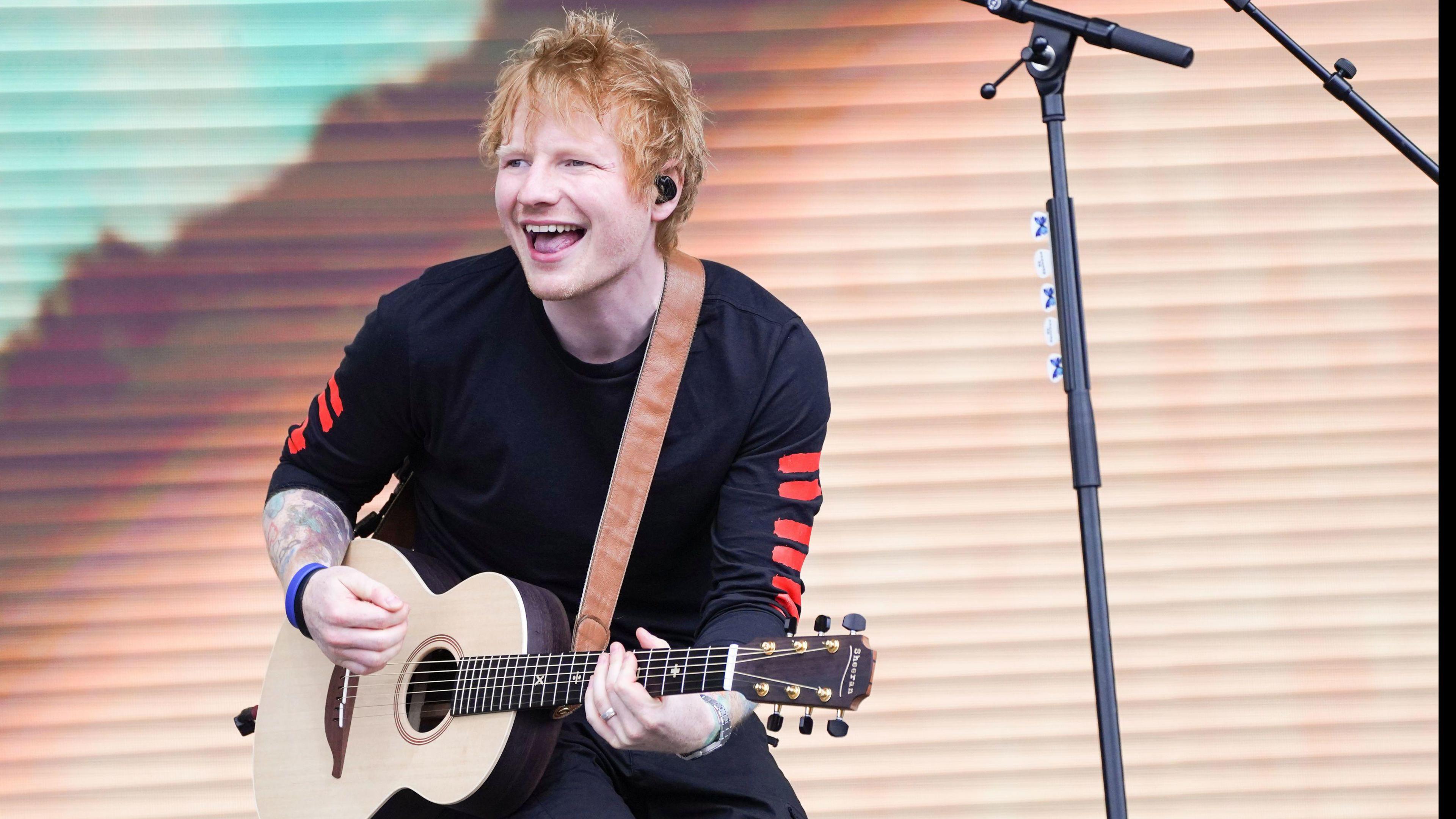 Ed Sheeran performing on stage during BBC Radio 1's Big Weekend in Coventry. He is smiling while strumming a guitar. He is wearing a black long-sleeved t-shirt with red stripes down the arms. A screen can be seen behind him.