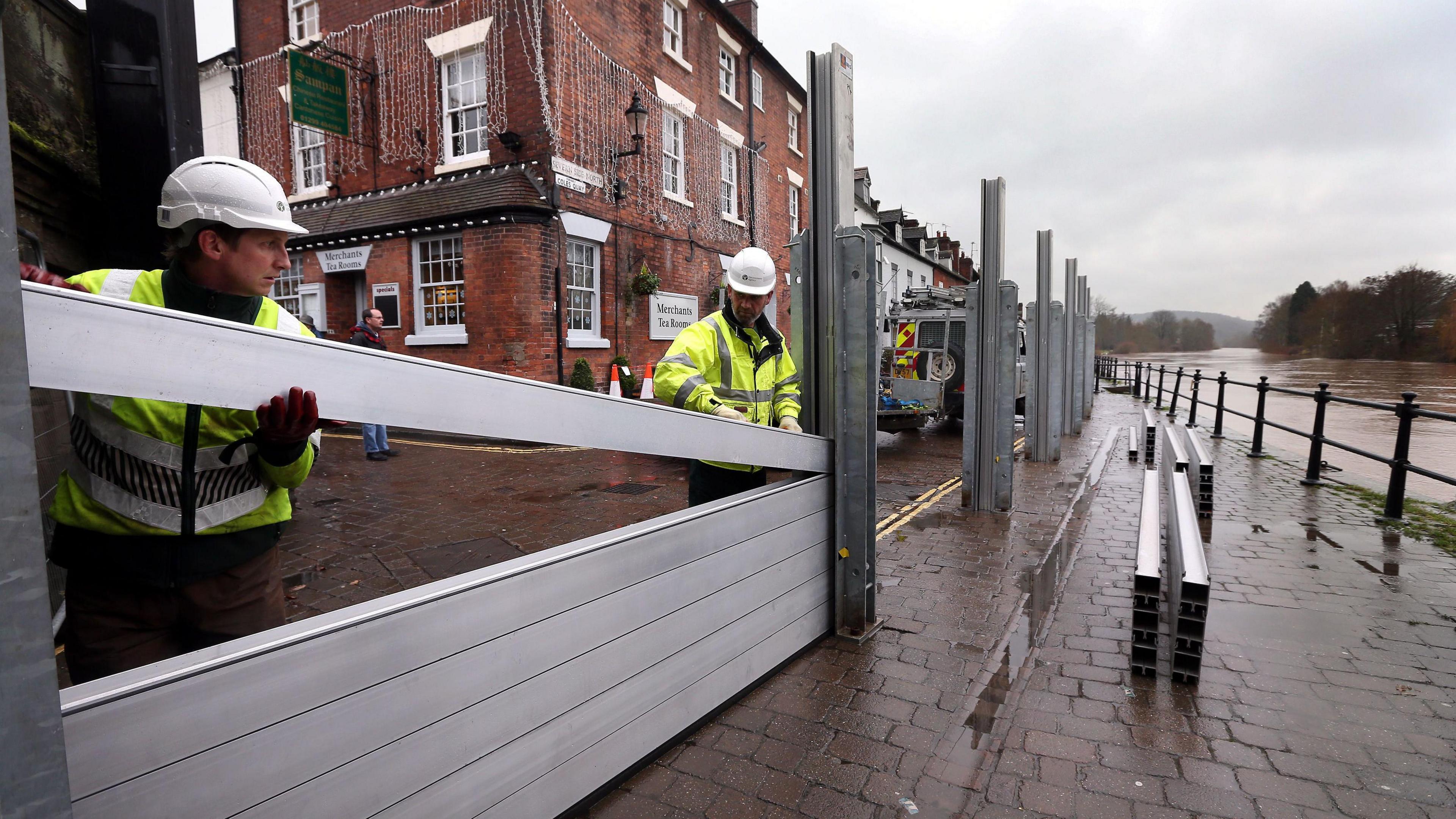 Two men wearing high-vis clothing and white hard hats slot metal beams into place, along the waterfront in Bewdley. The swollen River Severn can be seen on right, in front of the barrier, with a red-brick building behind the barrier that has white-framed windows, a green sign hanging on it, and decorative lights hanging from it.