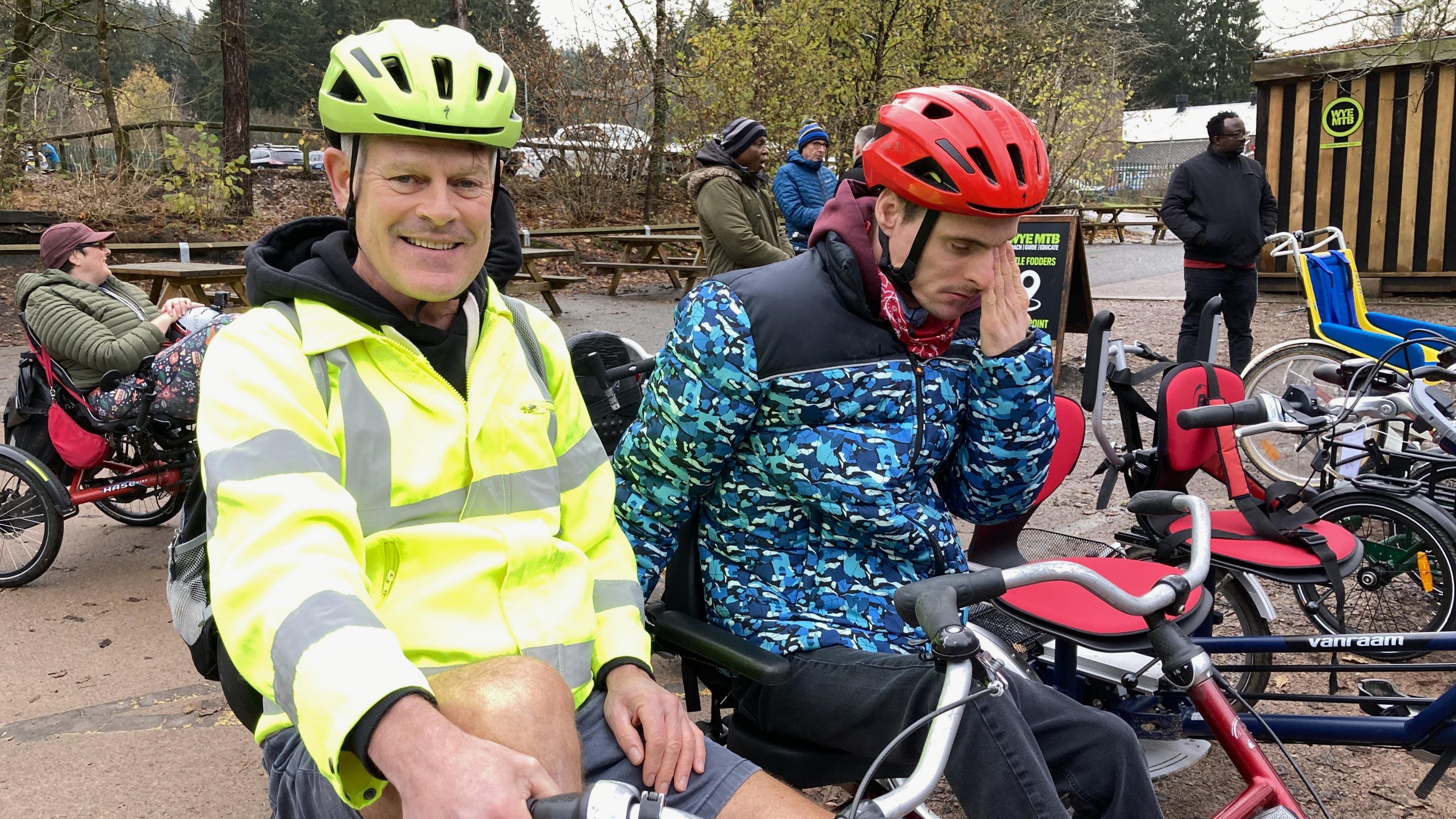 Zade Luker smiling at the camera, wearing a yellow high-vis jacket and yellow helmet. Simon has a red helmet and a teal camouflage jacket on. They are both sitting on a side-by-side bike and in the background there are other participants with their bikes.