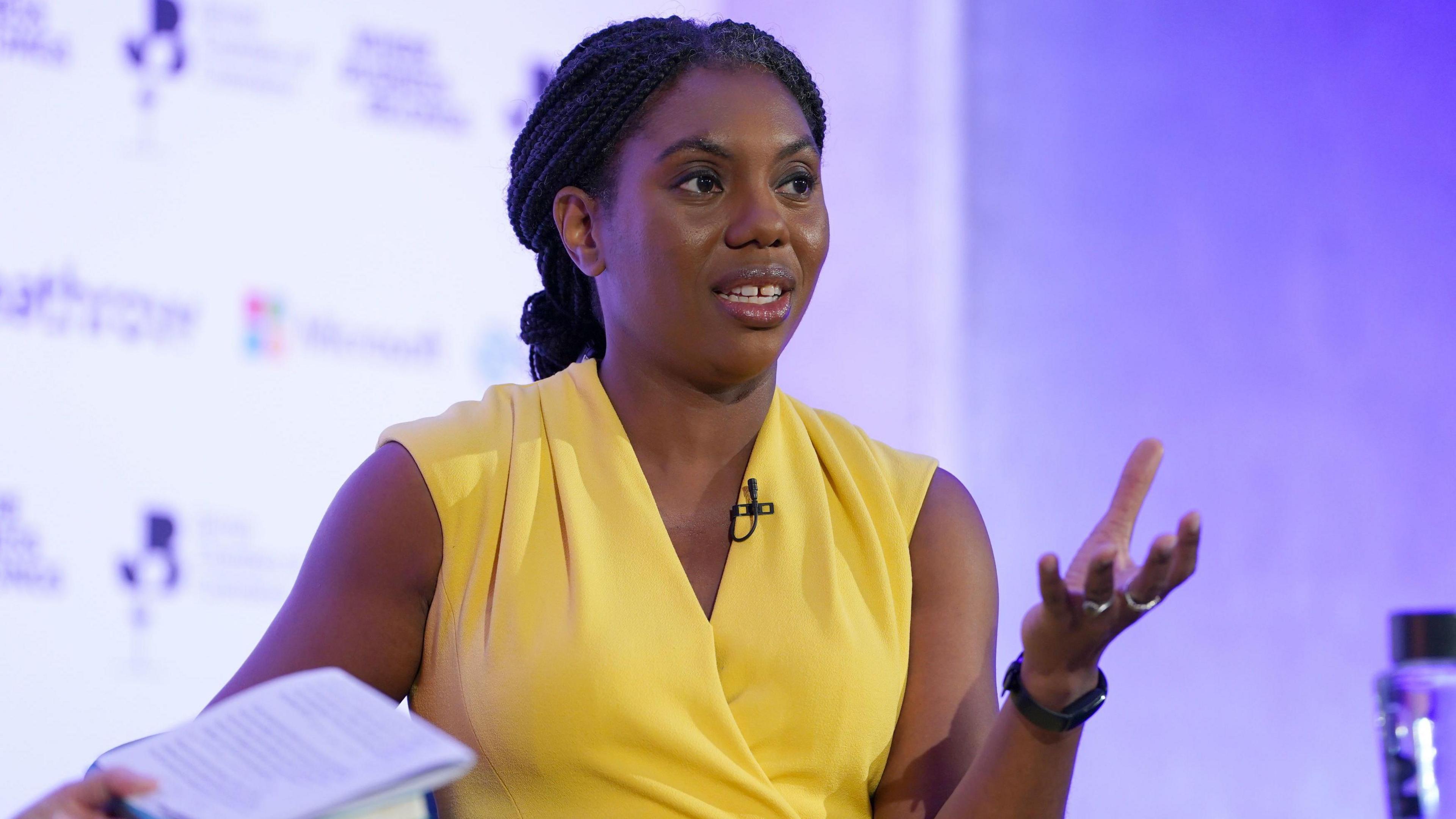 Kemi Badenoch wearing a yellow dress and holding a notepad