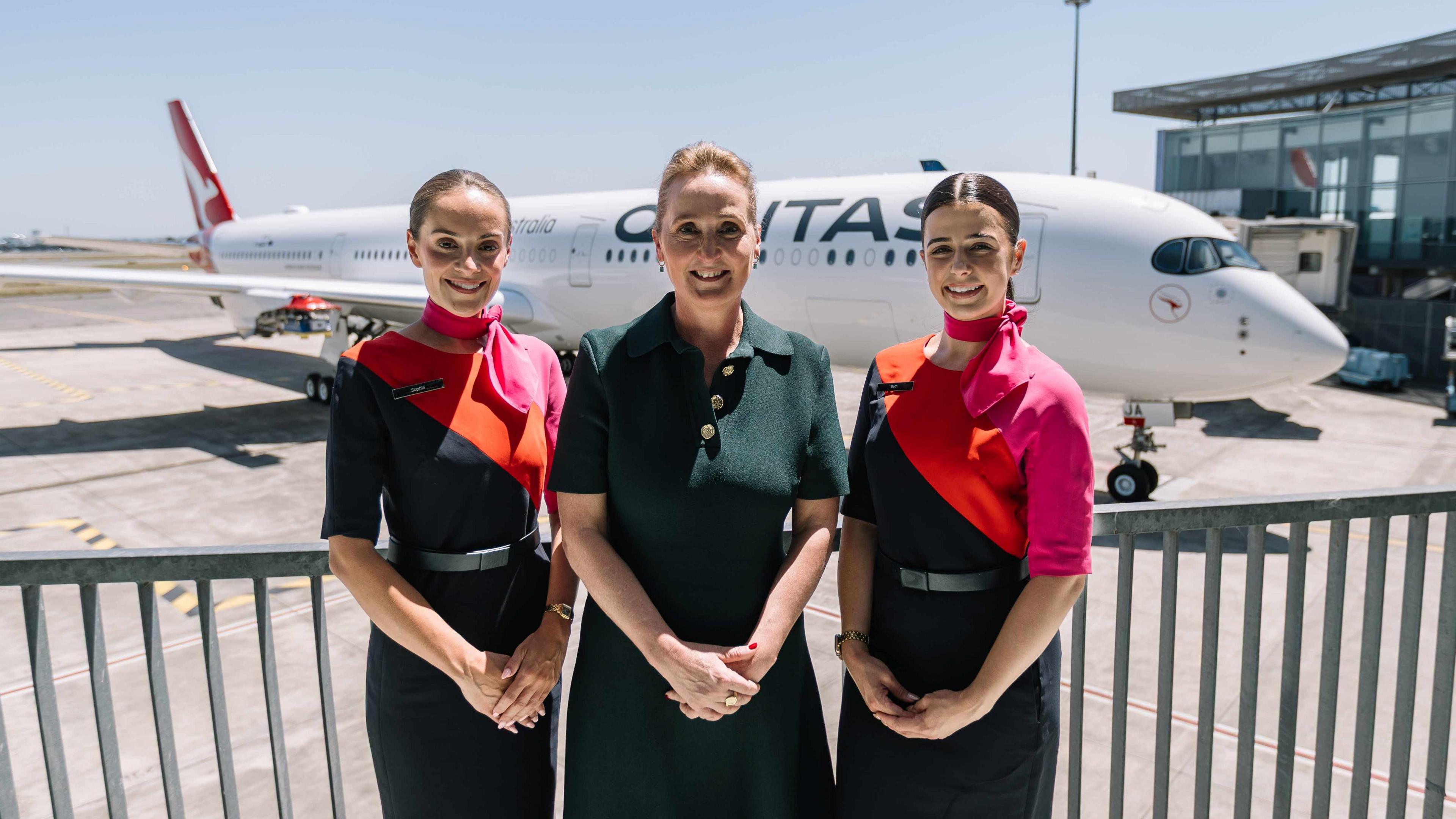 Three woman stand with their hands clasped in front of them in front of a large aircraft with 'Qantas' painted down the fuselage.