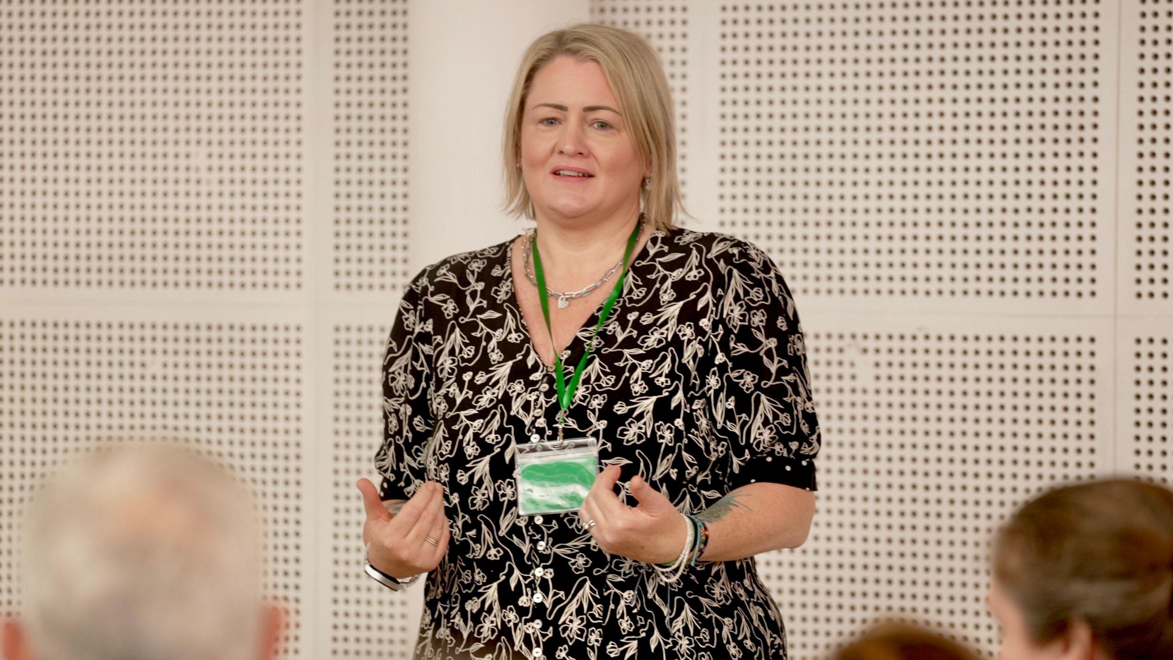Fiona Wells, a woman with a blonde bob, wearing a black dress with a gold pattern, standing in front of an audience as she addresses a meeting of the adoptive parents group she founded.