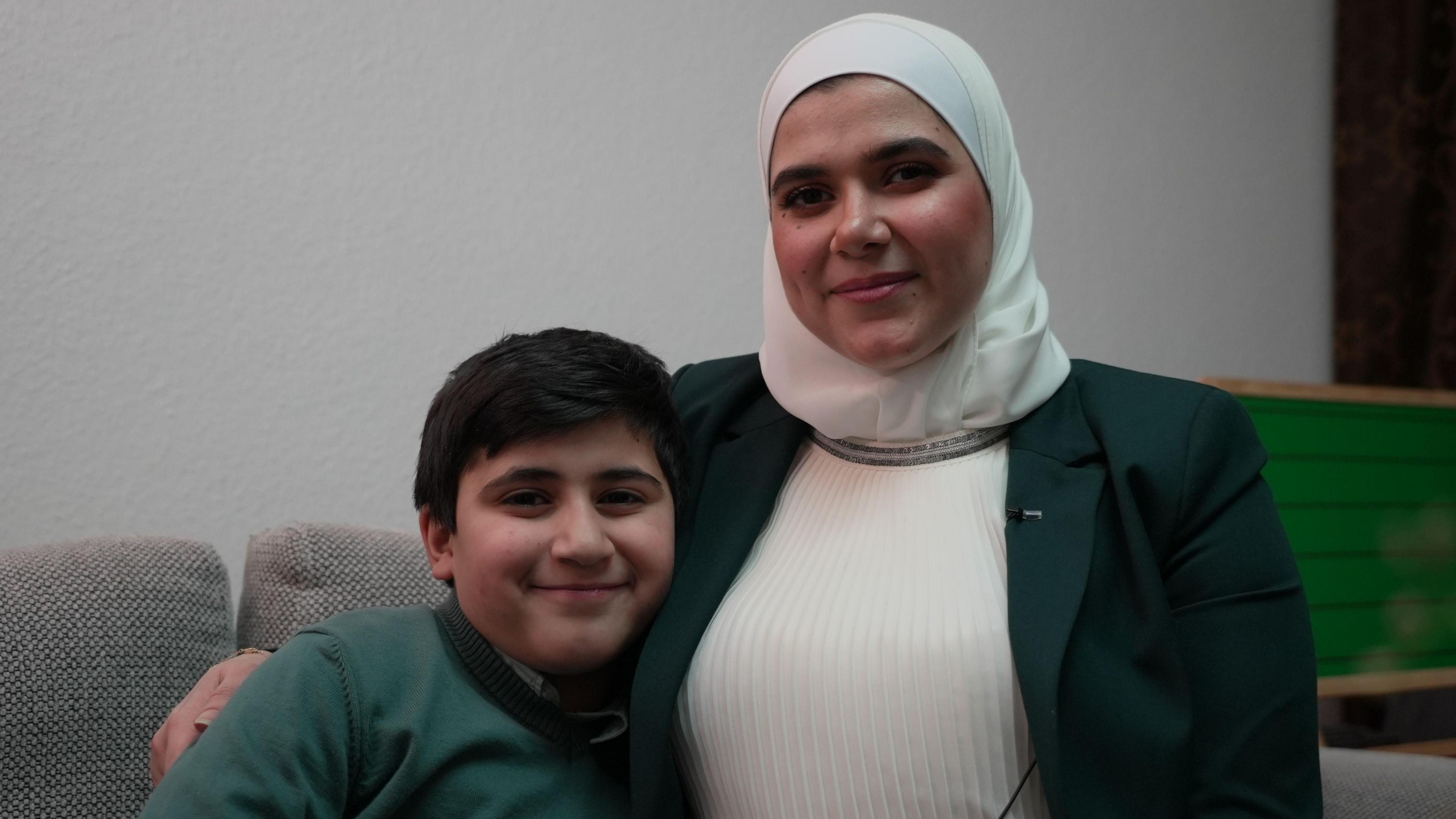 Alya and Rami pictured together. Alya, on the right, is wearing a green blazer and a white headscarf. Rami, on the left, is smiling at the camera and wearing a green jumper.