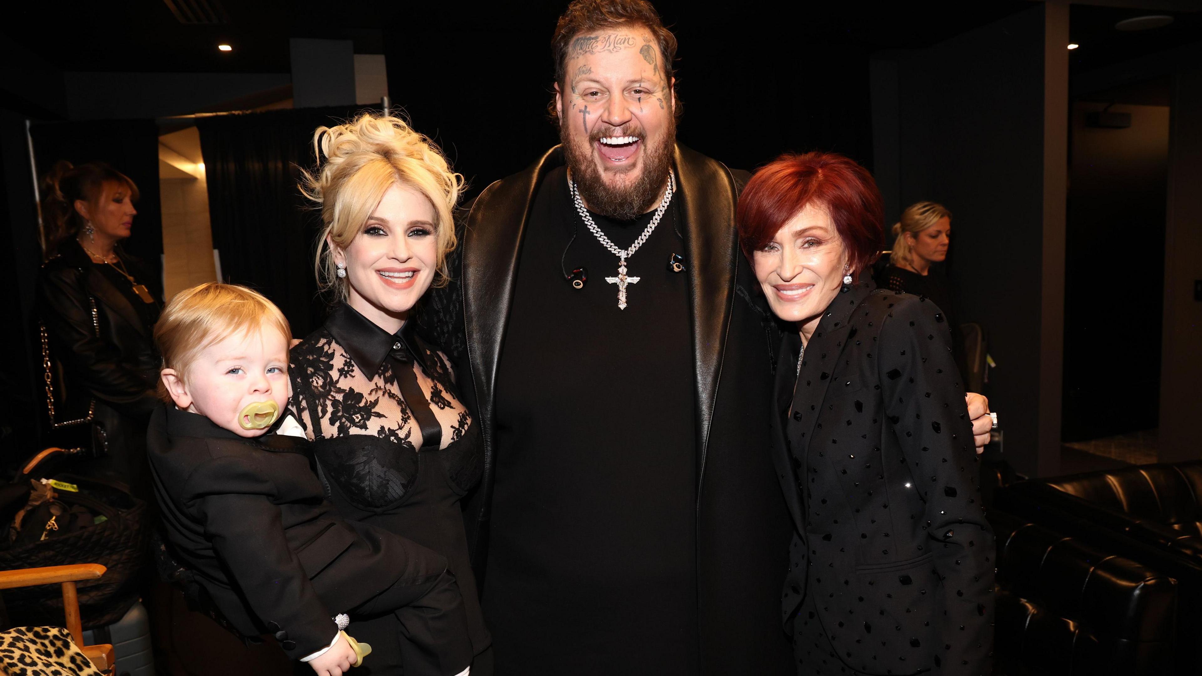 Ozzy's daughter Kelly has blonde hair and is wearing a black lace dress. She is holding her toddler son Sidney. A man, Jelly Roll, is wearing a leather jacket and a crucifix. He is smiling, has tattoos on his face and has short brown hair and a beard. He has his arm around a woman, Sharon Osbourne, who has red hair and is wearing a black coat. She is smiling.