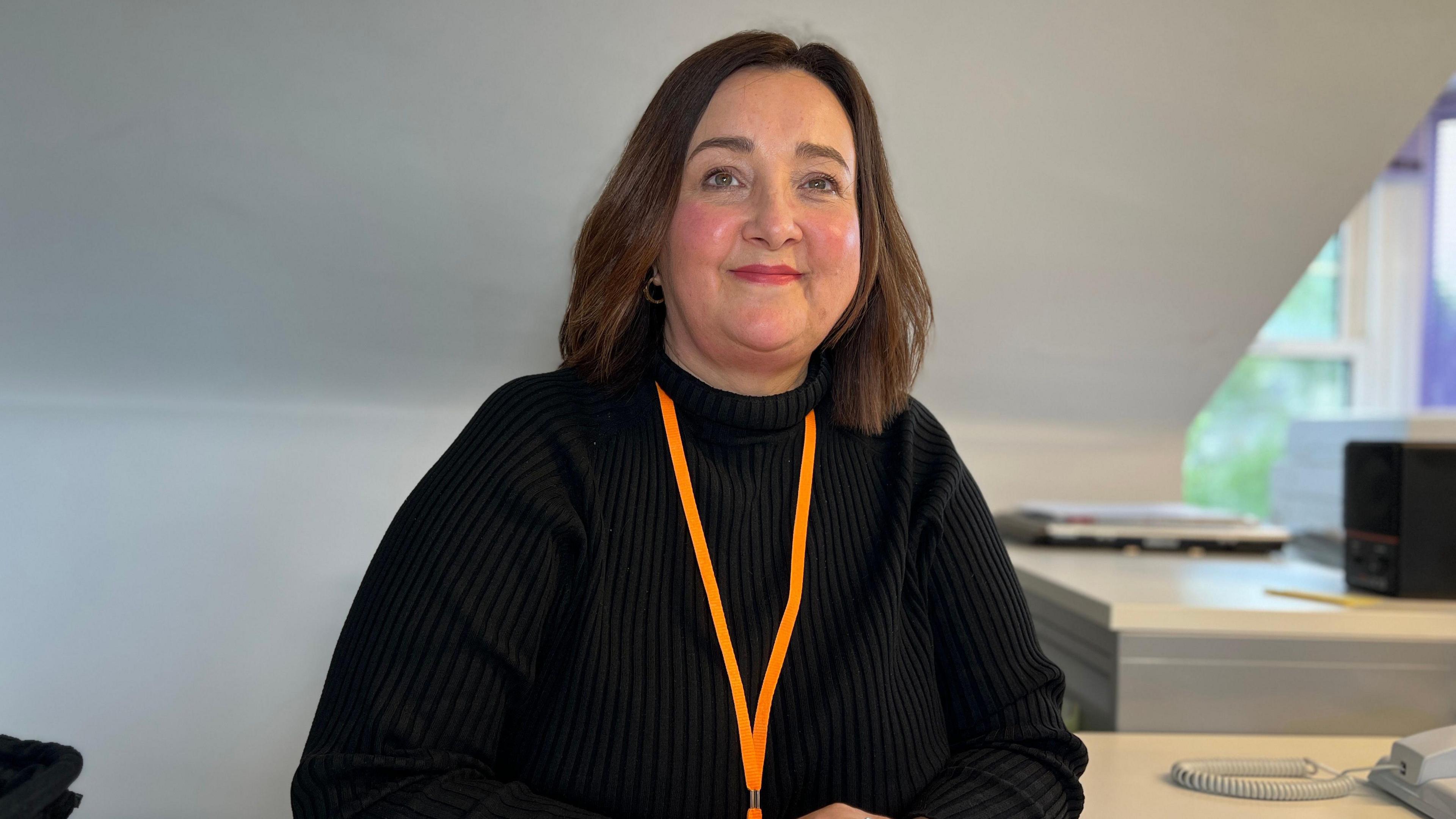 Fiona smiles at the camera as she sits at a desk with a landline phone next to her