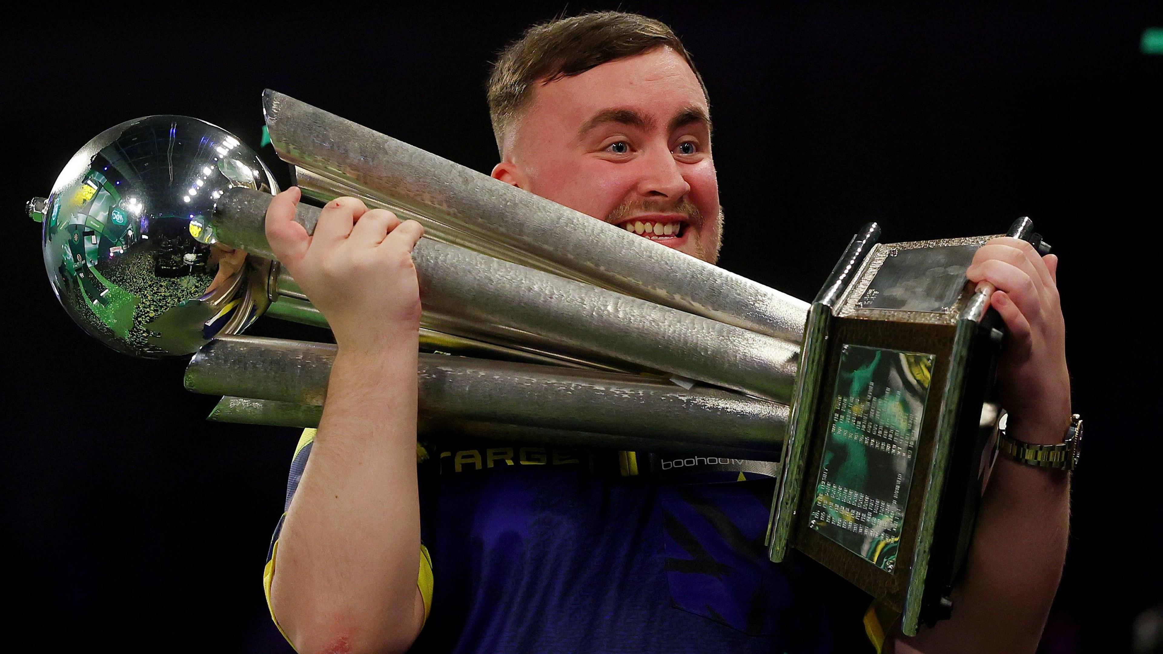 Luke Littler has a smile on his face as he lifts up the large Sid Waddell Trophy
