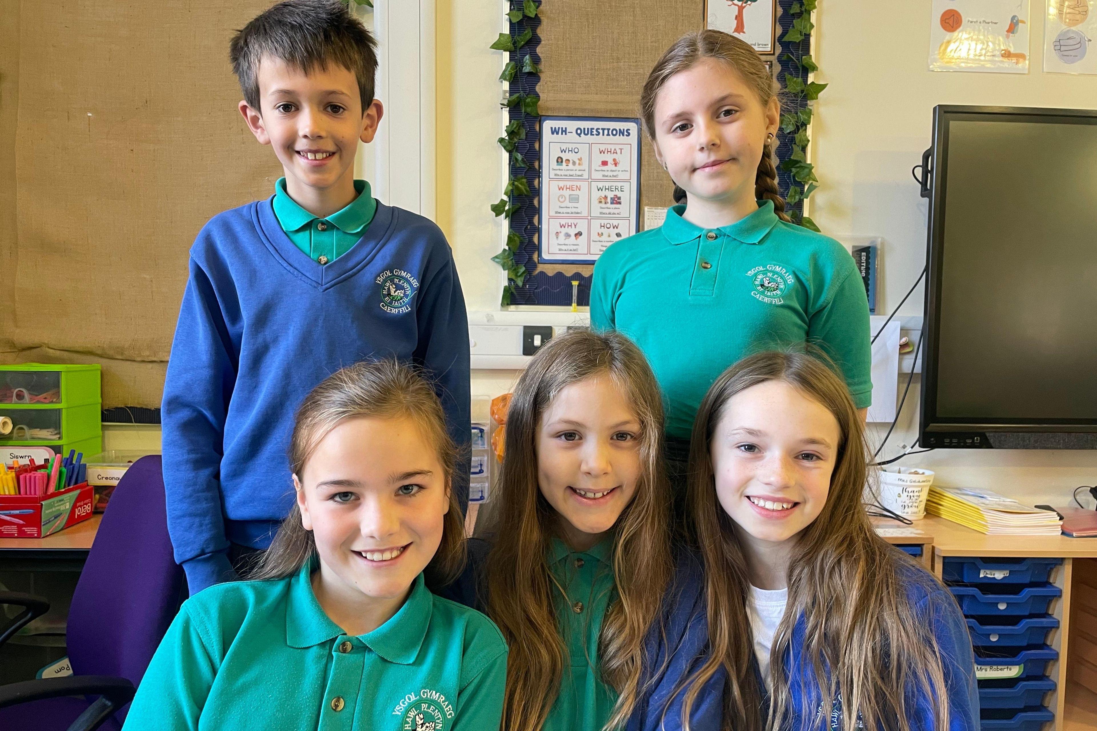 Pum plentyn mewn gwis ysgol glas a gwyrdd gyda bachgen a merch yn sefyll yn y cefn a thair merch yn eistedd o'u blaenau.
