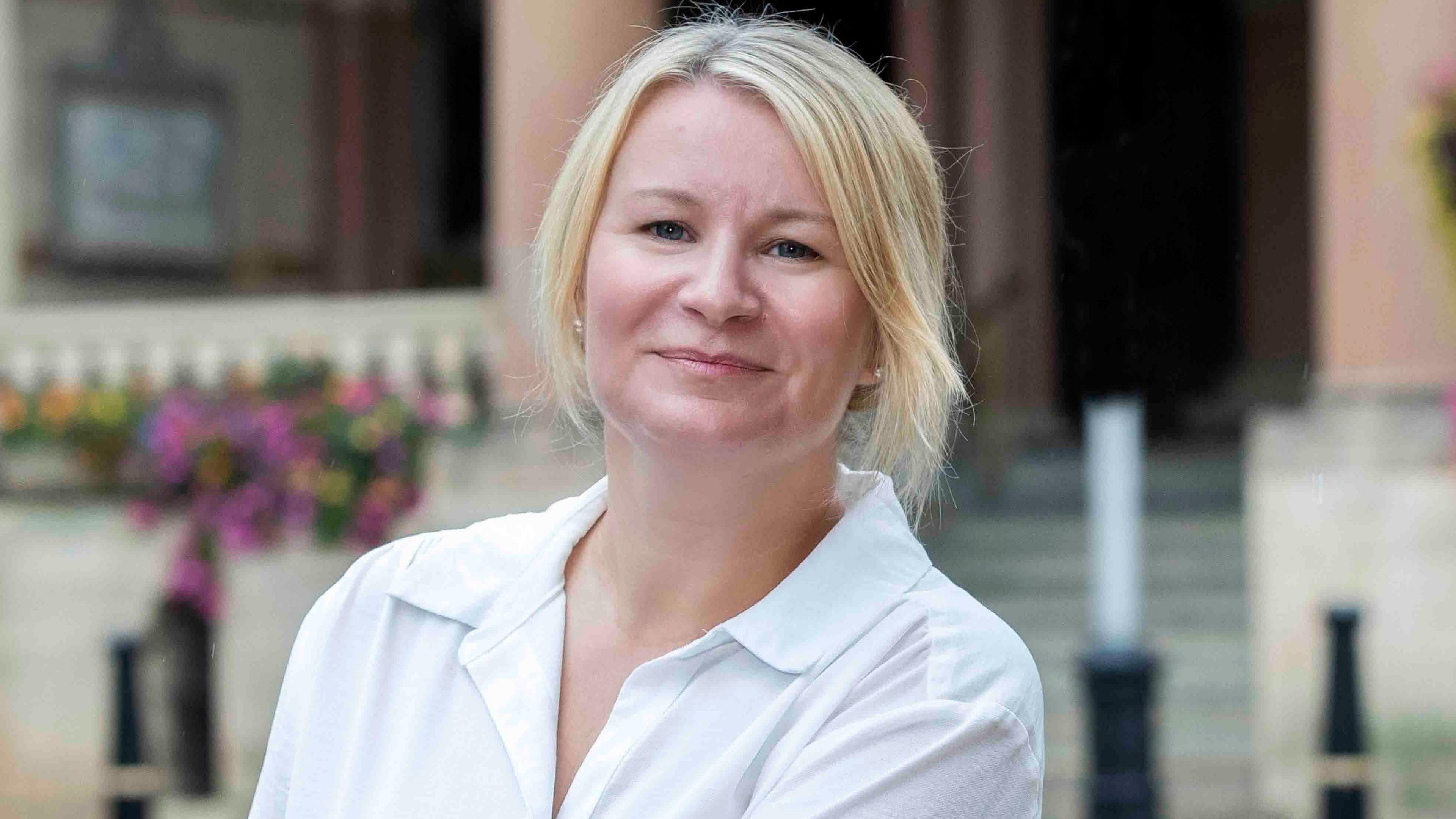 Sarah Ward, with long blonde hair, tied back, wearing a white shirt, looking at the camera and smiling, outside a building, which is blurred in the background, with flowers to the left. She has earrings in both ears.