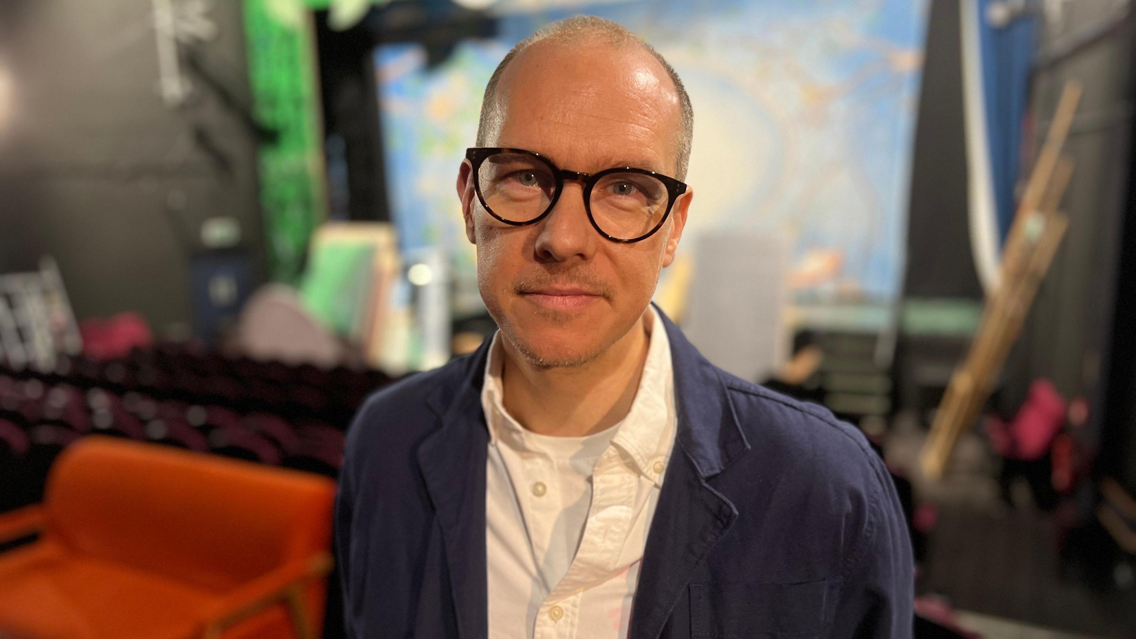Derek Bond is standing in the middle of the picture with a blue jacket and white shirt - he has glasses and in the background is stage with theatrical props and furntiture.