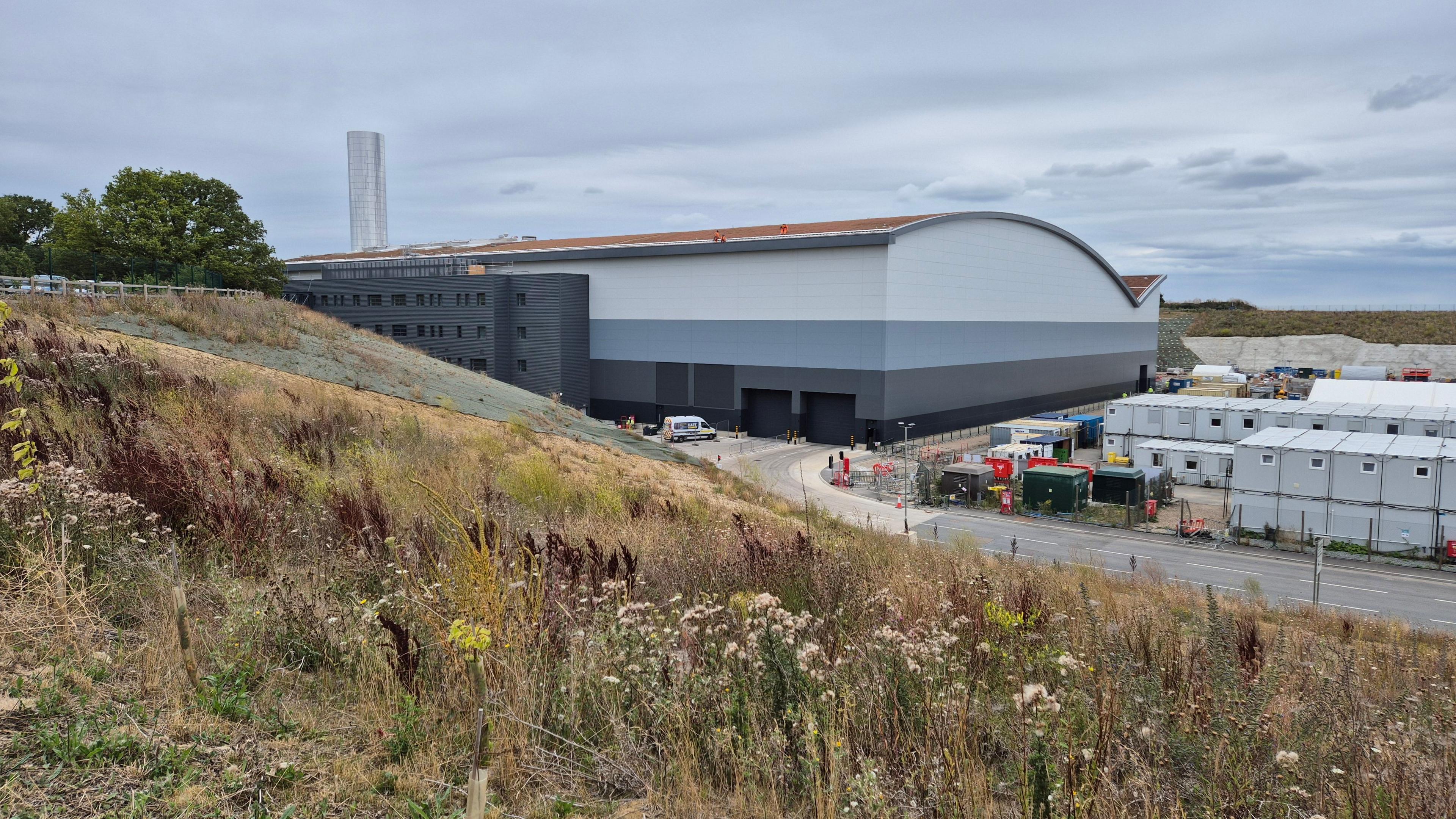 The Integrated Waste Management Facility (IWMF) at Rivenhall, which is an incinerator designed to burn Essex County Council's household waste. It has a domed roof to the incinerator building, covered with browned grass. Next to it are several large pipes, and a 35-metre tall chimney which is covered in a reflective surface. The building is set 15 metres into the ground, and a large slope leads down from the left-hand side of the frame down to the base level. There are several grey modular buildings next to the main incinerator building, as well as an access road.
