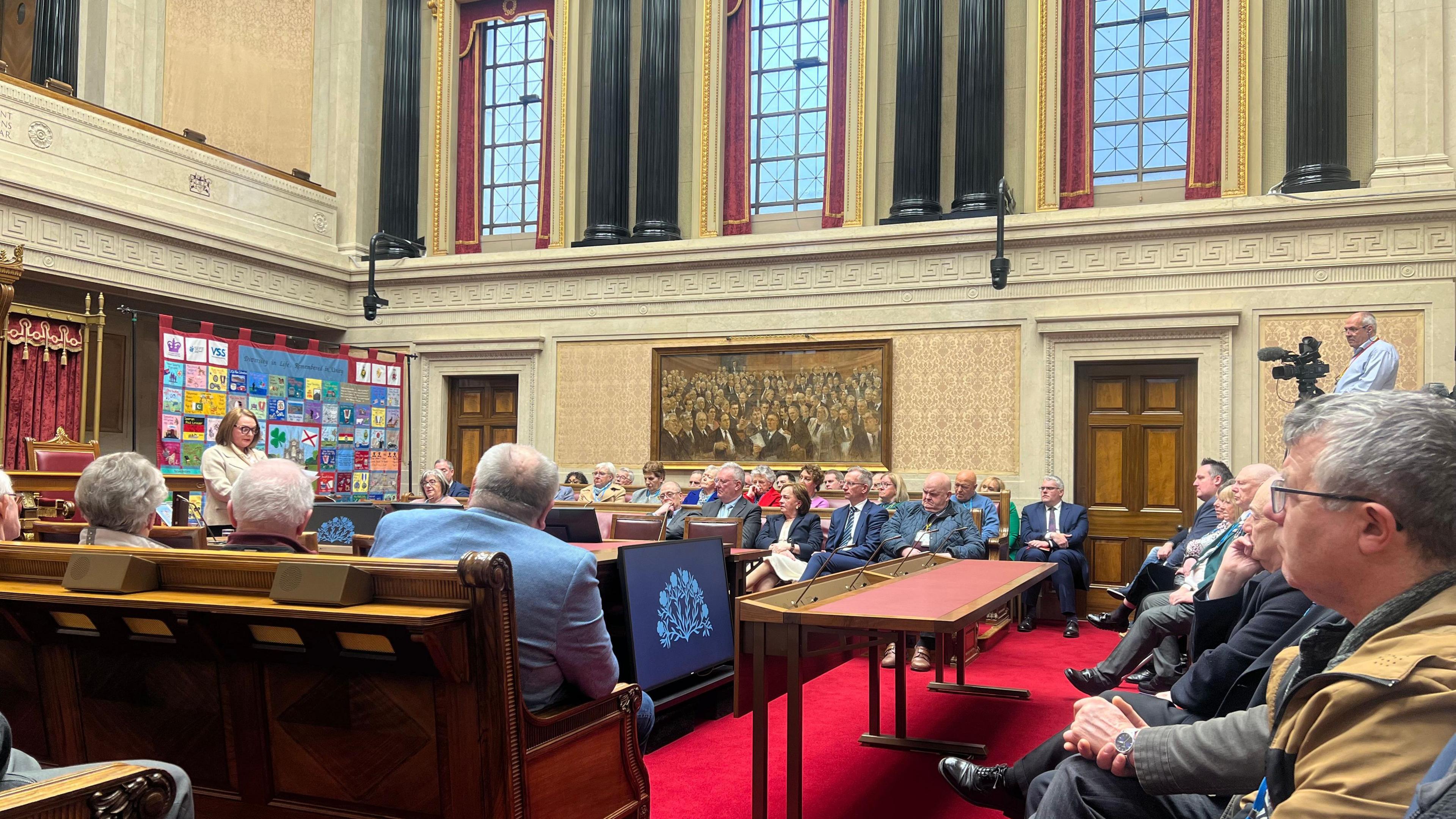 Caroline D'Eath speaks at Stormont. There are chairs around the room and she stands in front of a patchwork memorial.