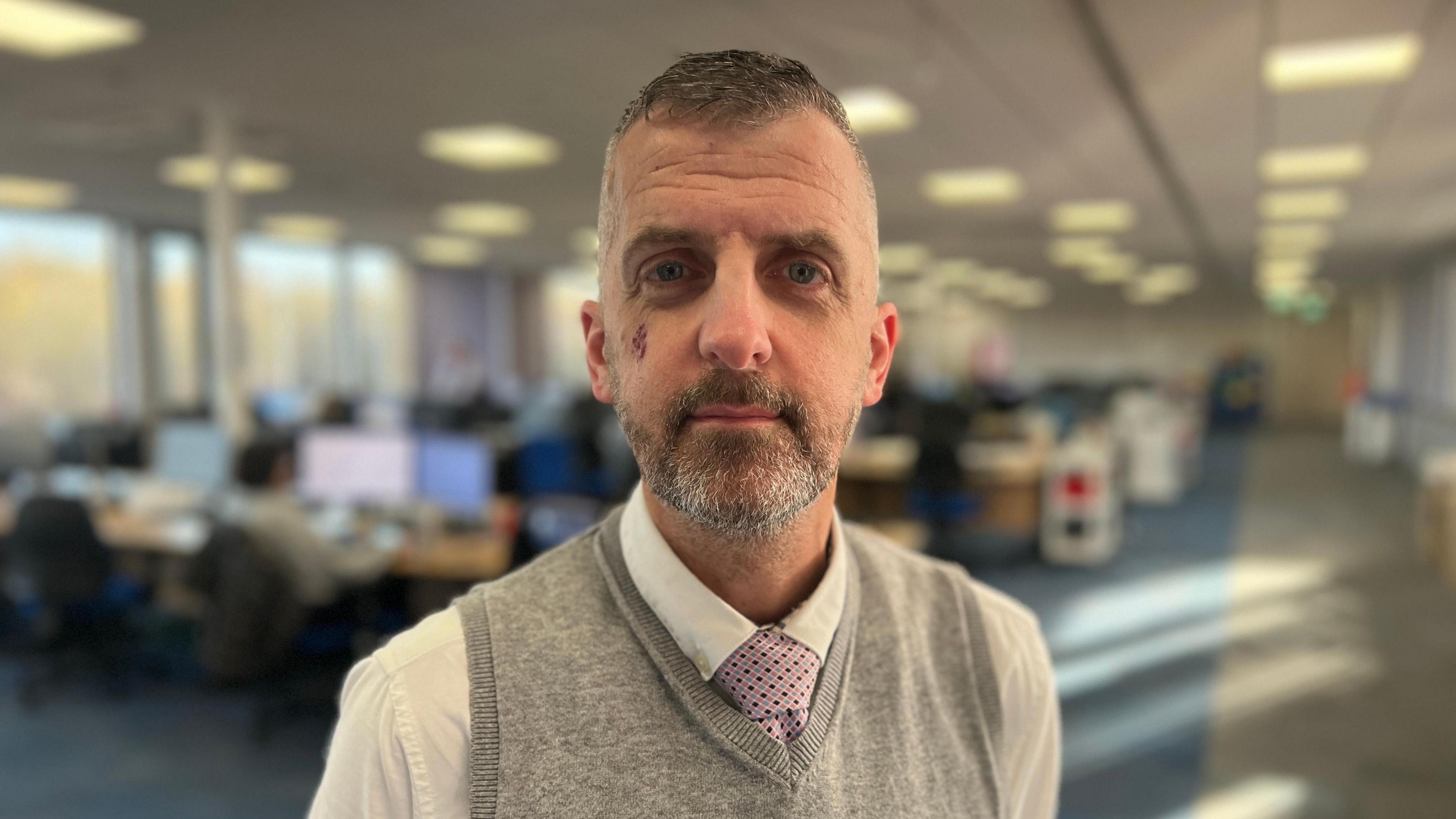 Christian O'Brien looking into the camera with a blurred out office behind him. He has short grey hair and a beard, and is wearing a grey sleeveless jumper, white shirt and purple tie.