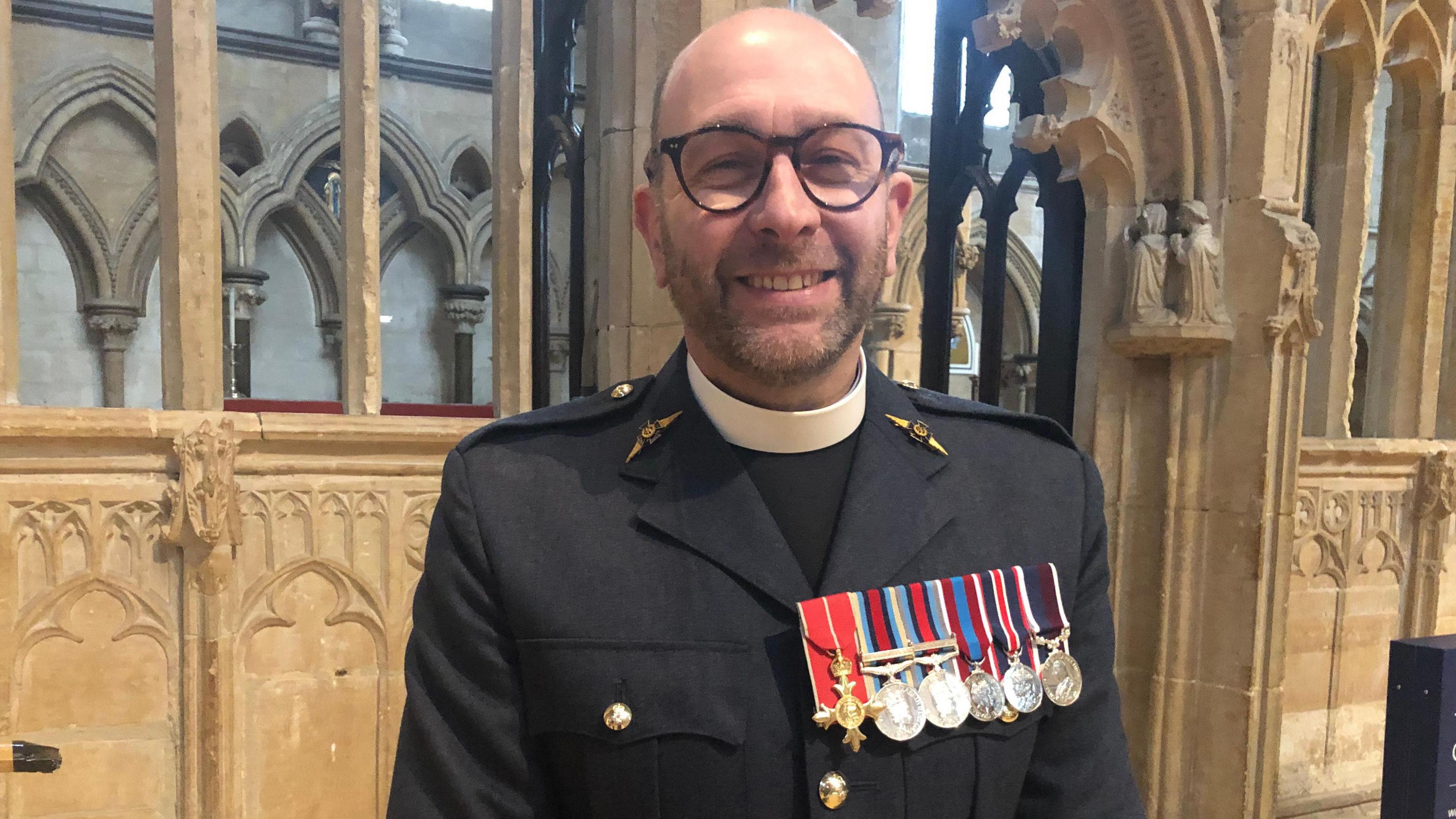 A bearded man stands in front of stonework at Lincoln Cathedral, wearing a blue RAF uniform, medals, a clerical collar and glasses.