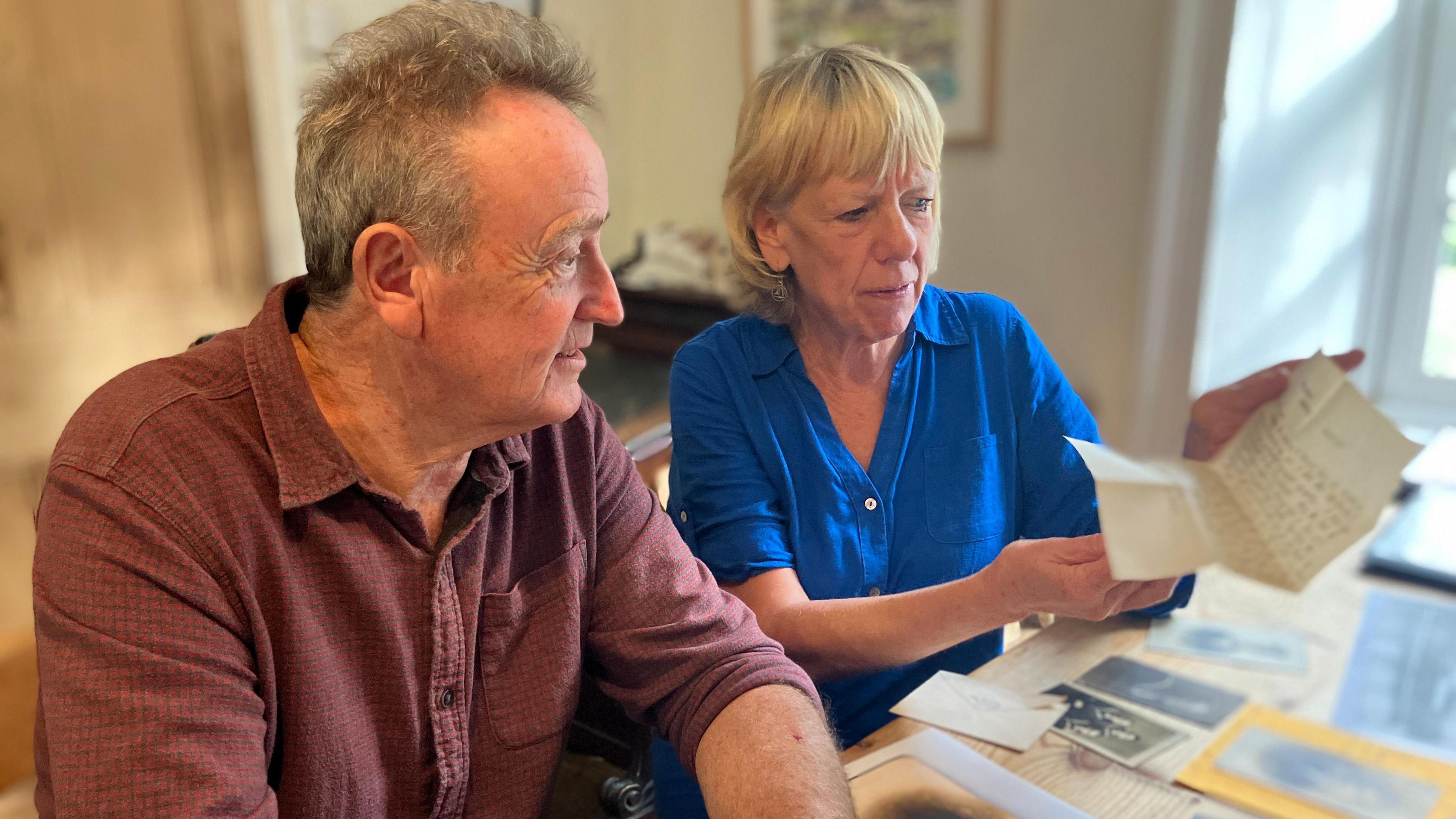 Mike and Henrietta Sandford sit at a wooden table covered in black and white photos. Henrietta is holding a handwritten letter and showing it to Mike