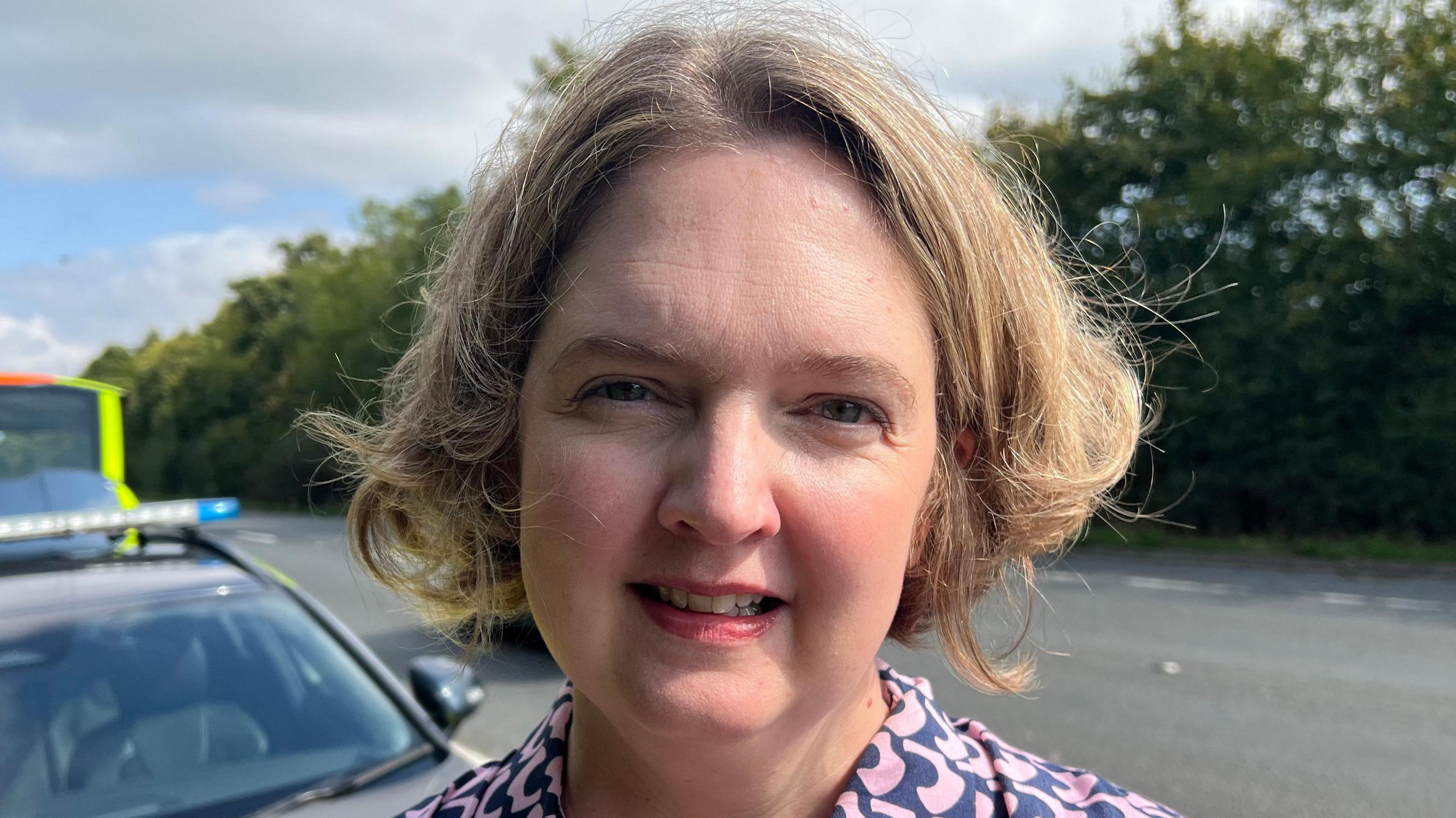 Local MP, Anna Sabine. She has short blonde hair and is standing beside the road, looking at the camera. She is wearing a blue and pink patterned collared shirt.