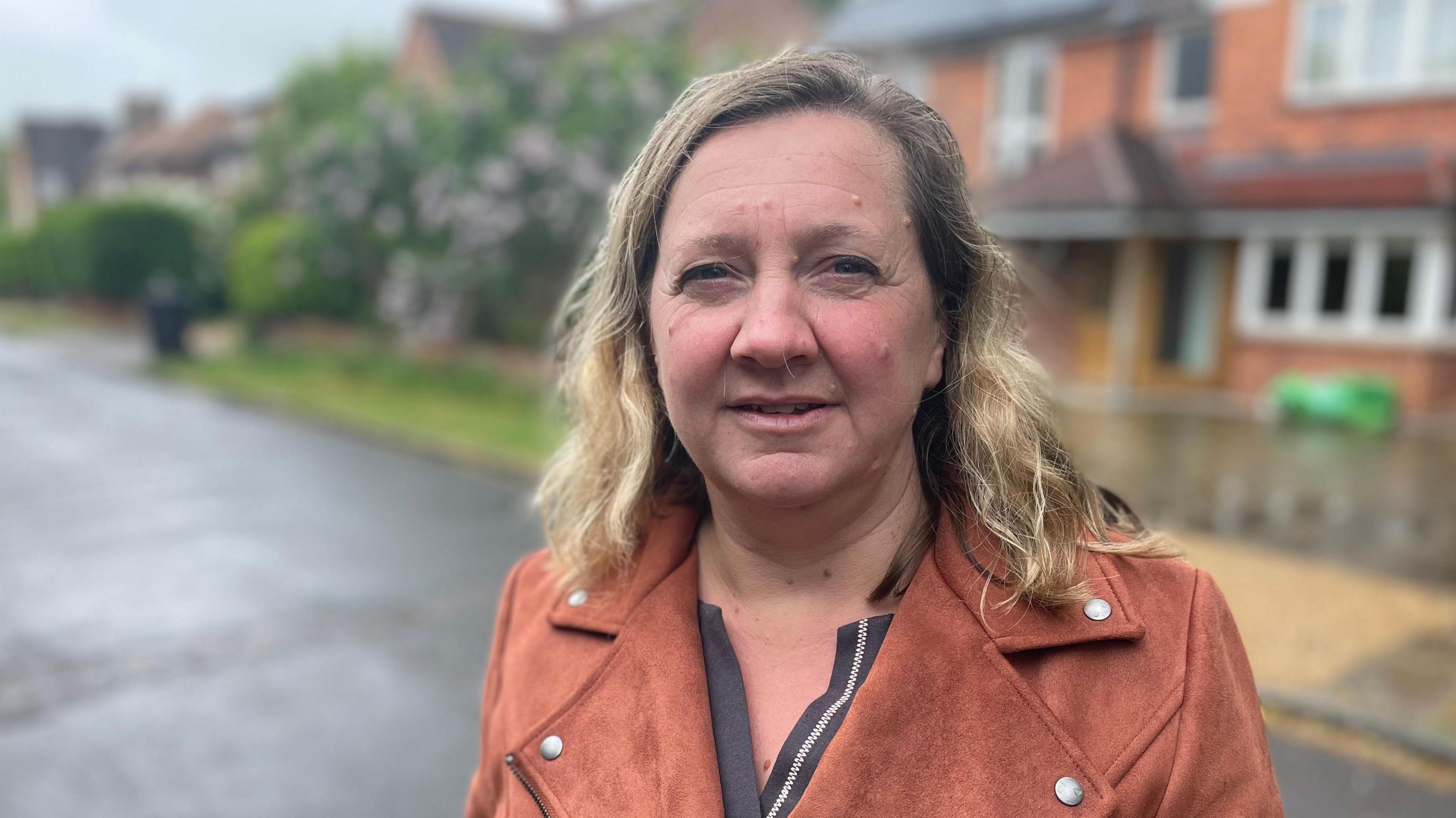 Lucy Nethsingha, wearing a butterscotch coloured coat over a grey v-necked top. She has shoulder-length fair hair and is looking towards the camera. She is standing on a street with blurred out houses behind her