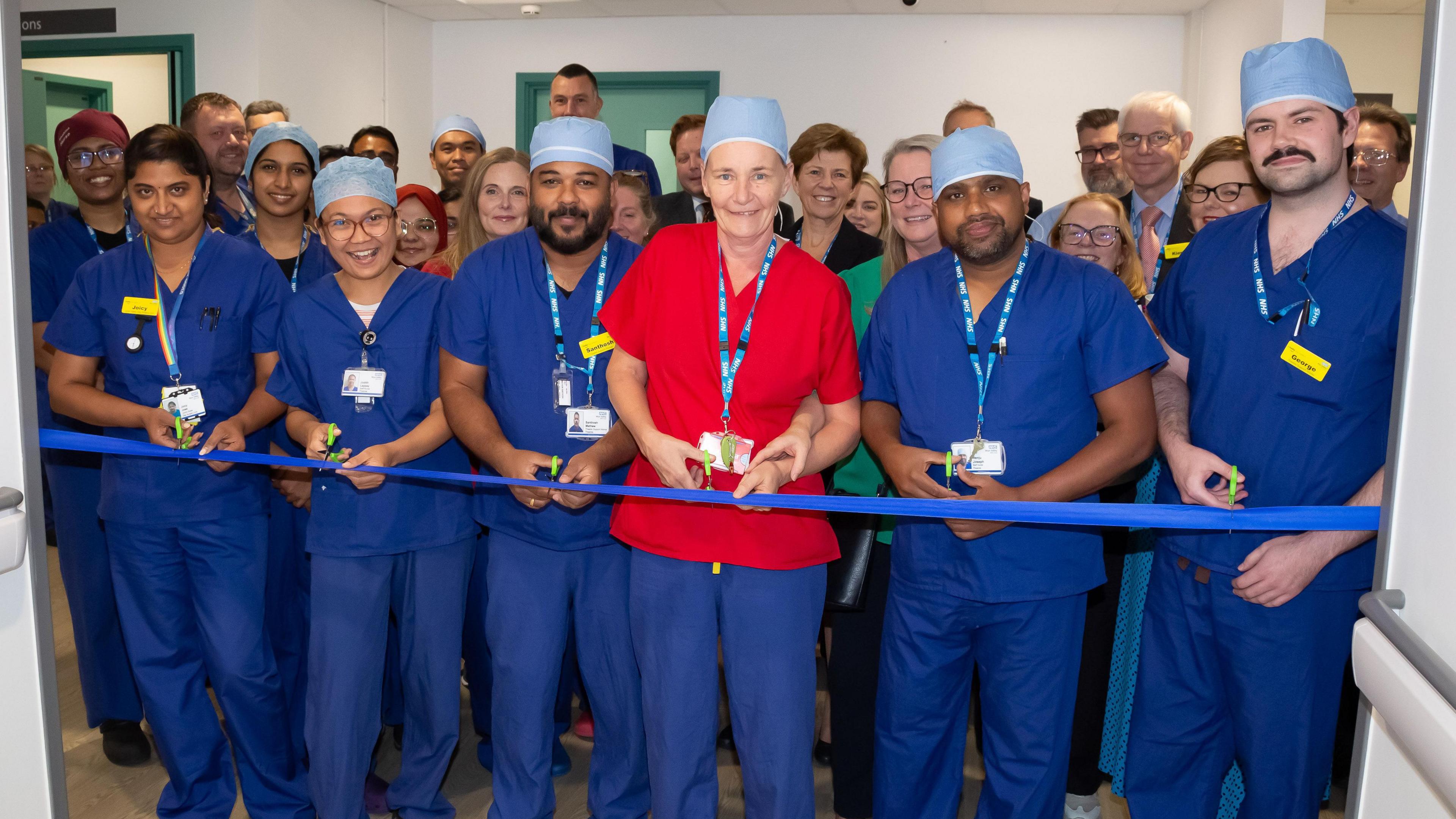 Theatre staff dressed in blue scrubs working in the new Daycase Surgical Unit. A woman in red scrubs is cutting the ribbon to declare the new facility open 