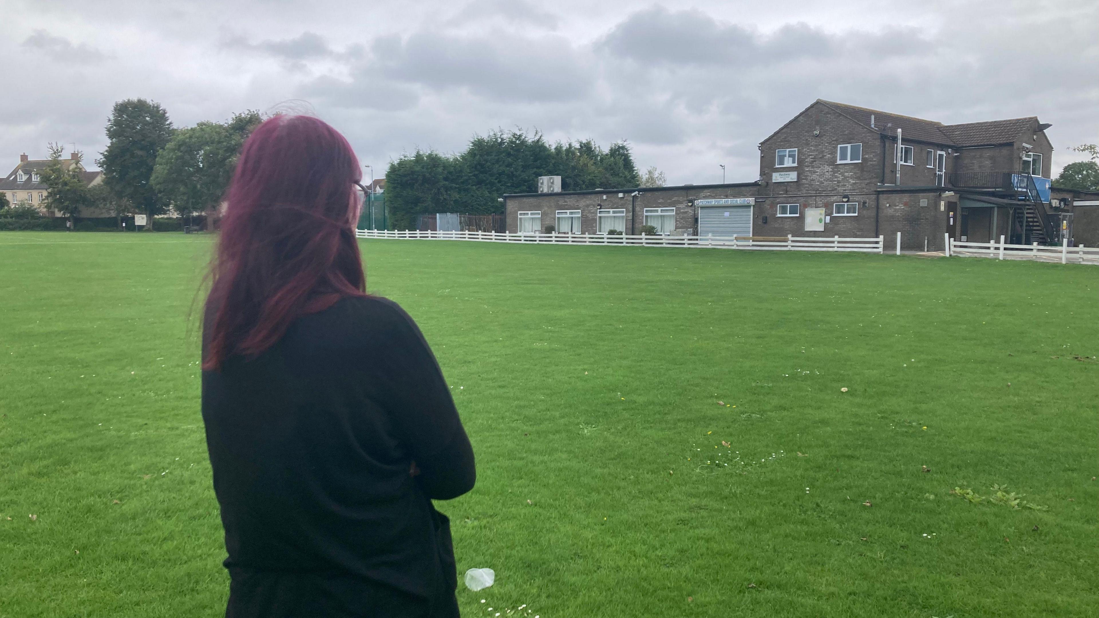 The back of a woman with dark purple hair wearing a black cardigan. She is standing on a green pitch looking towards the Patchway Sports and Social Club. It is a dark brown brick building with closed metal shutters. 