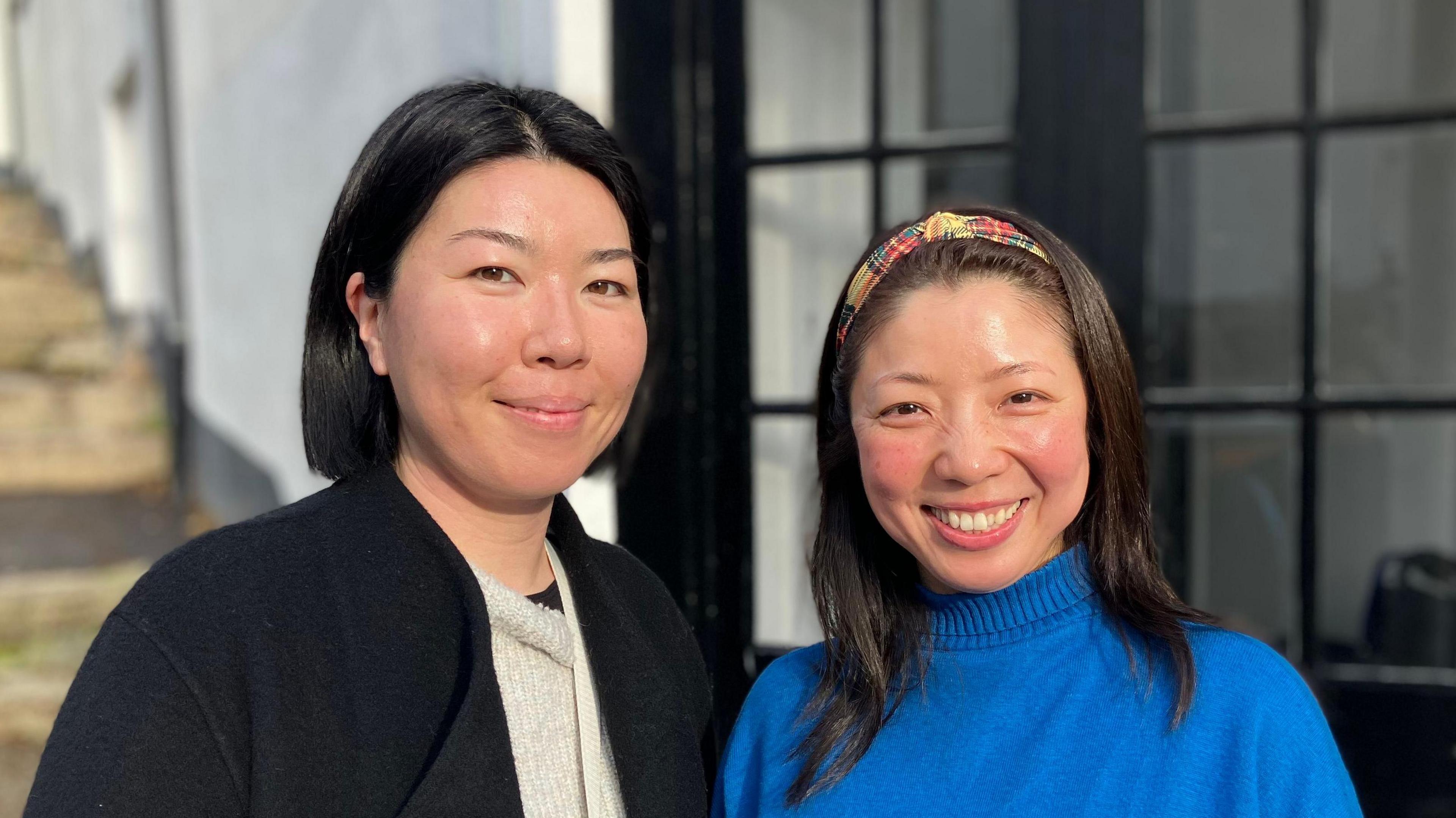 Two women stand in front of a traditional historic doorway in St Ives, Cornwall. One is wearing a black coat, the other a bright blue jumper. 