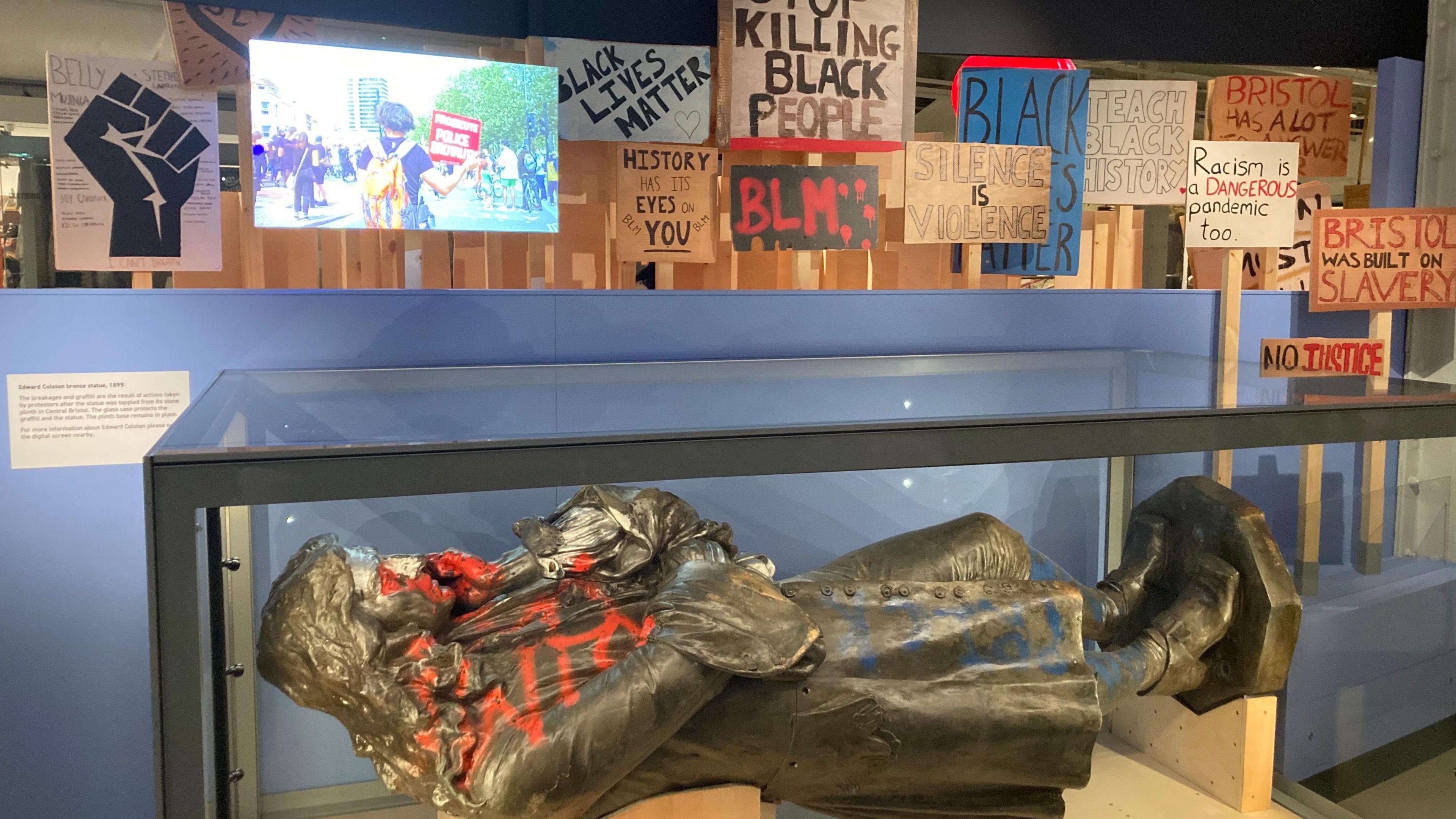 The Edward Colston statue lying on its back as part of the exhibition, with various Black Lives Matters placards in the background