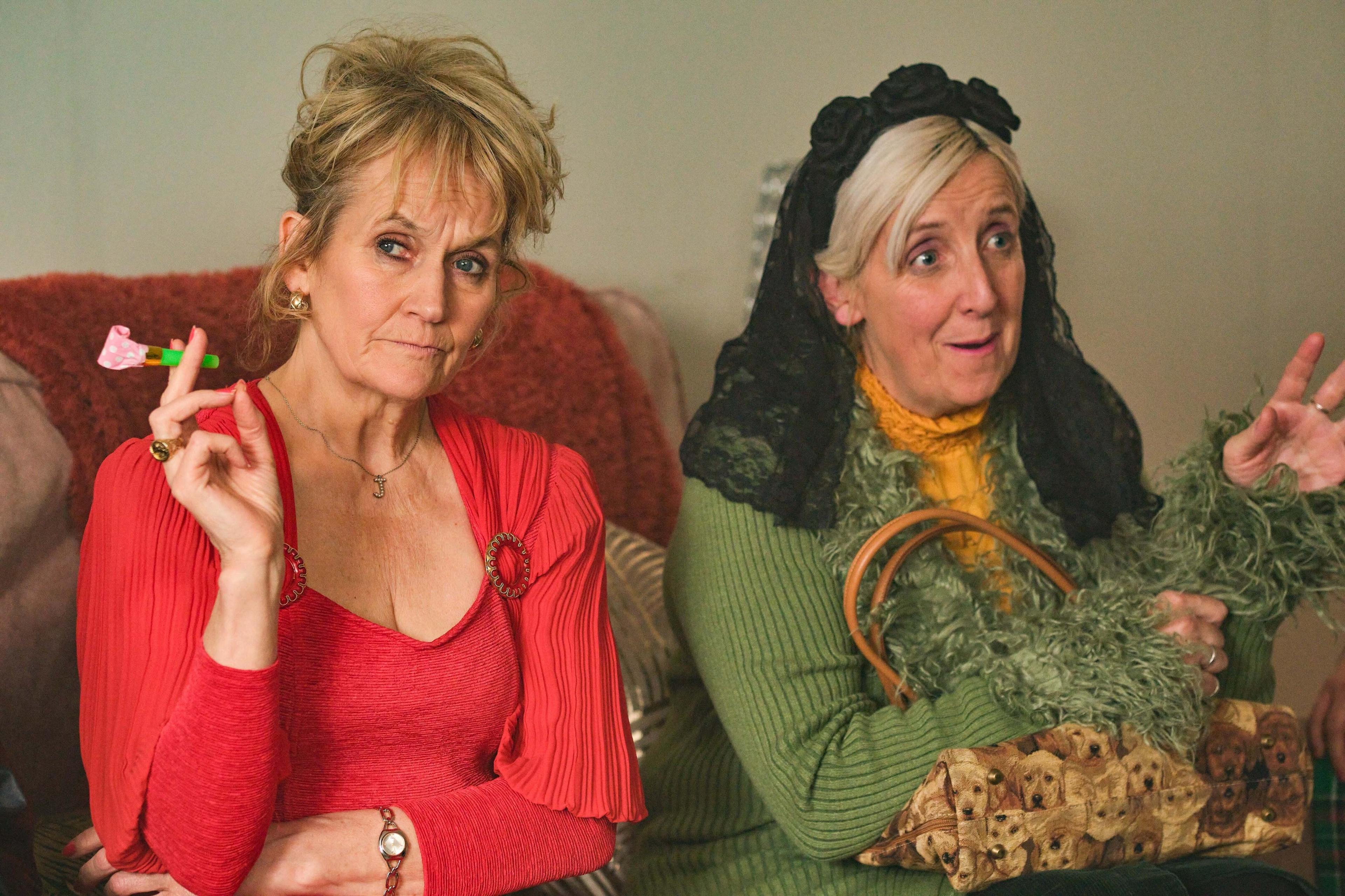 Lorraine Ashbourne (Joan) with Aunty Ange (Julie Hesmondhalgh) - Lorraine is wearing a red low-cut top and Ange has a black veil on her head and is wearing a yellow top and green cardigan