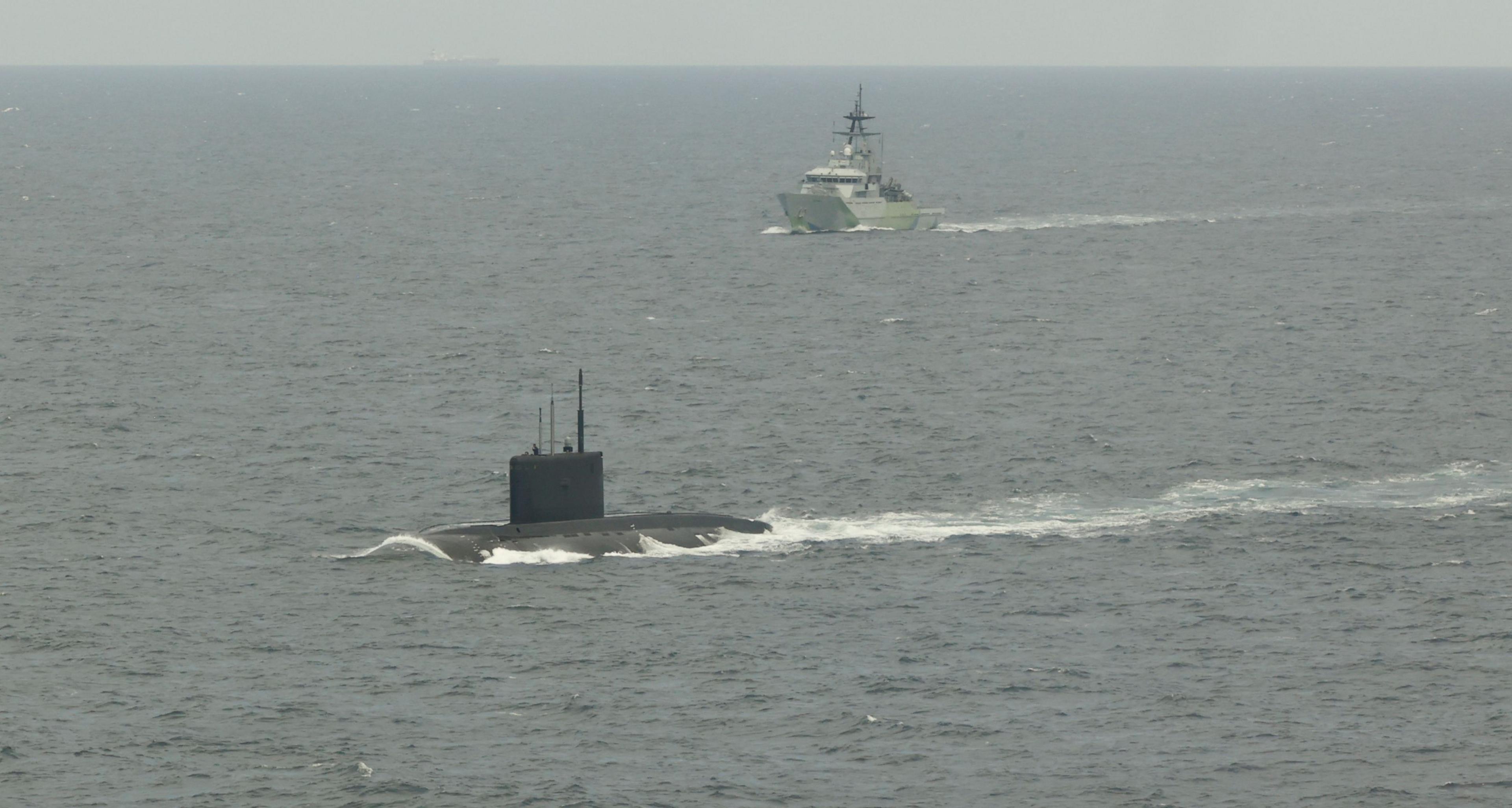 A submarine at the front of the image in the water with HMS Mersey in the distance