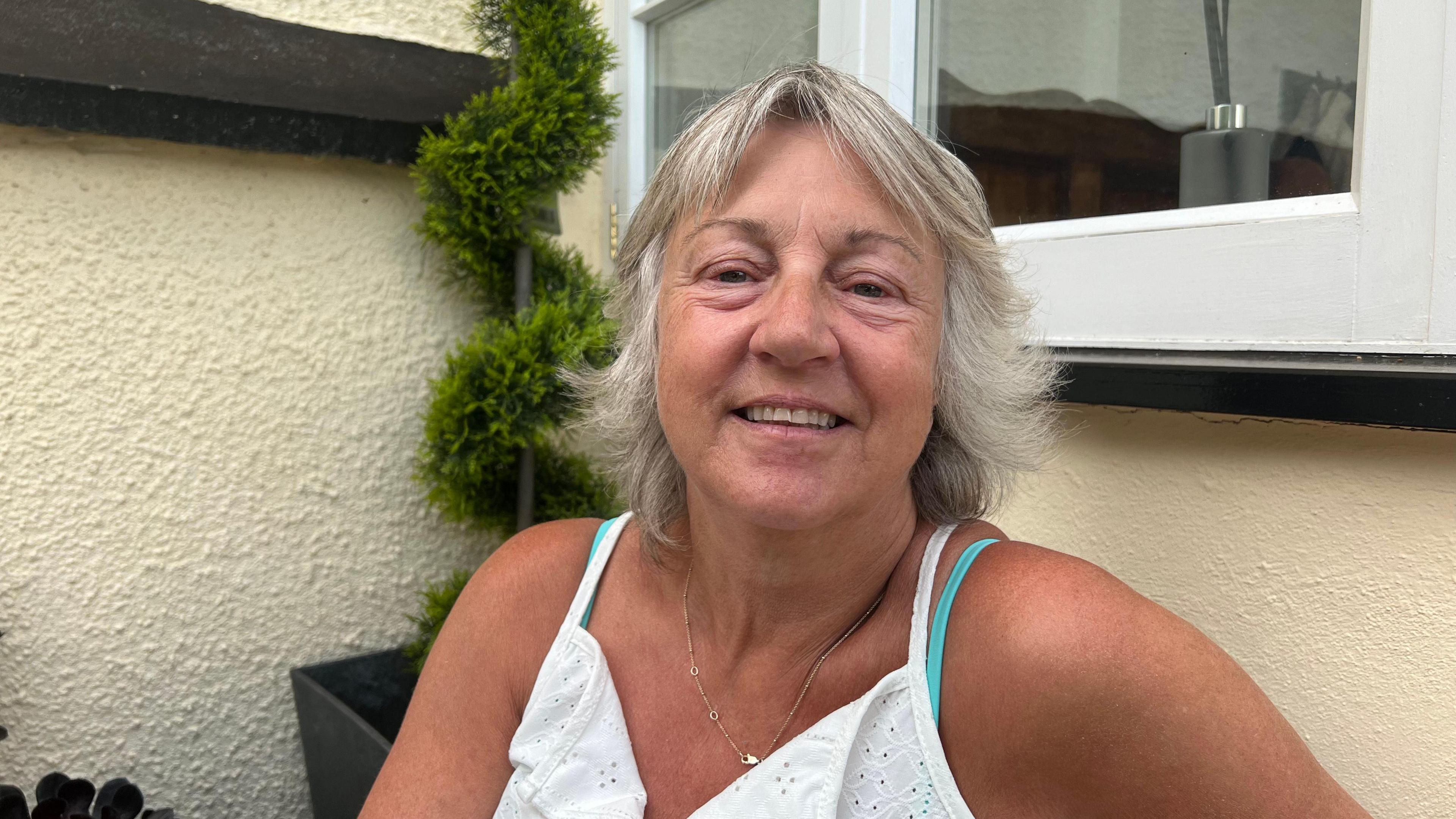 A woman in a white vest top with short blonde hair is sitting outside a cream house with a window. She is smiling at the camera.