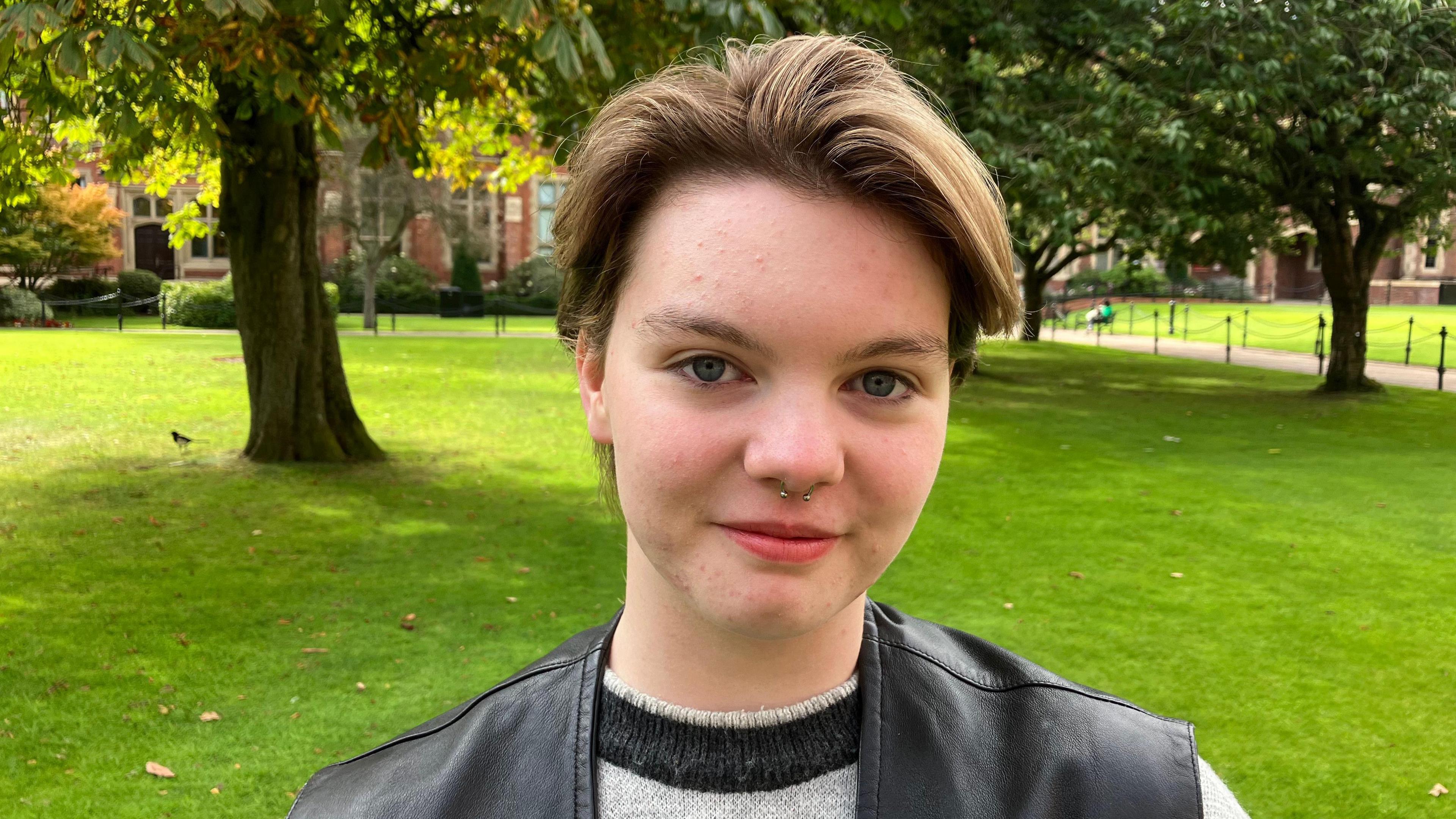 Matt is standing in front of trees in the shade.

They have brown hair pushed to the side and red lipstick on. 

They are wearing a black leather jacket and a grey jumper with a black turtle neck. 