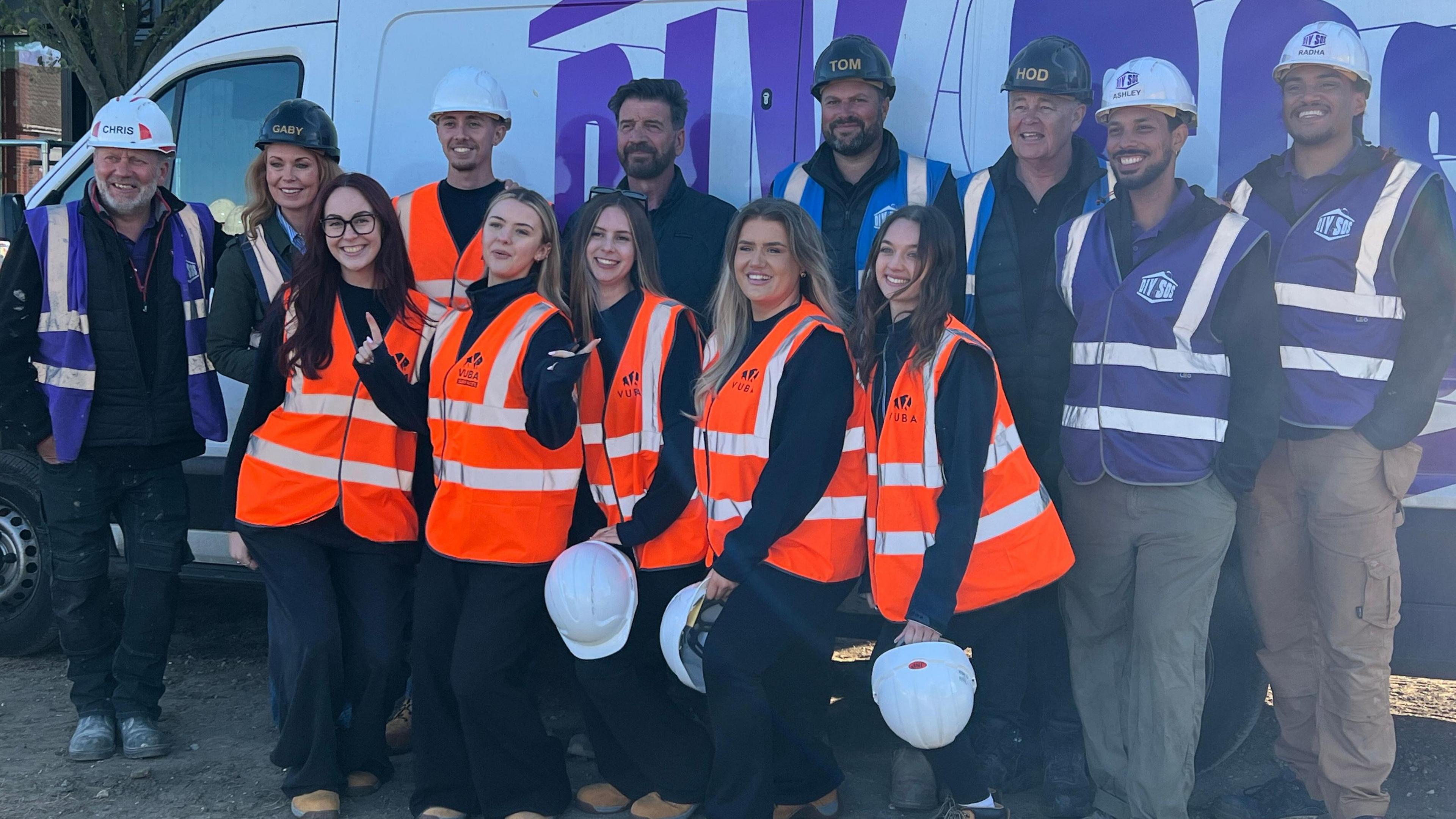 A group of people posing in front of a white van with "DIY SOS" branding. Some are wearing hard hats and purple or orange hi-visibility vests. They are smiling.