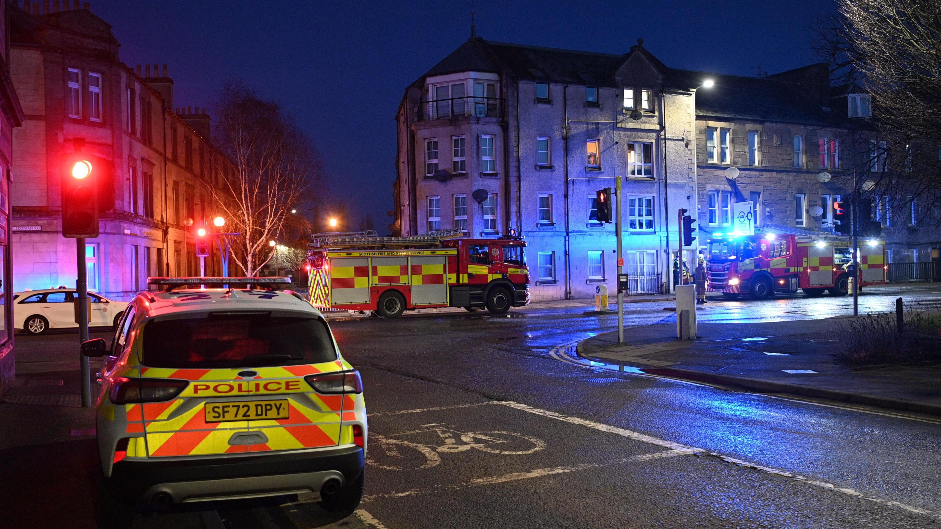 Two fire engines outside a four-storey block of flats.  A police car is in the foreground of the shot.