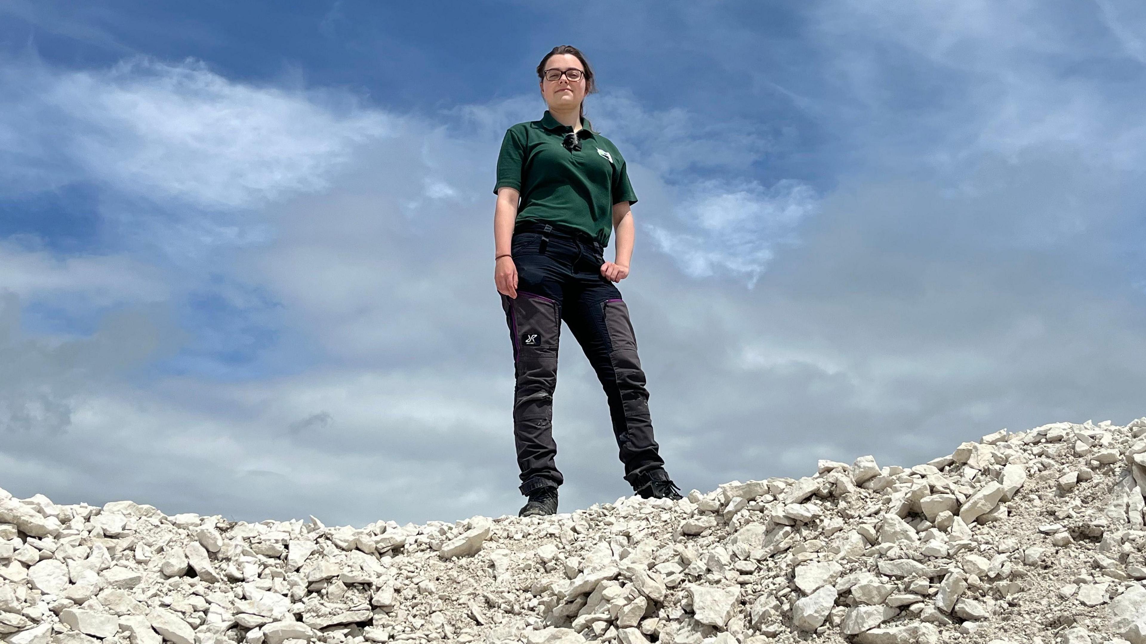 A woman in black trousers and green polo shirt stands on top of a pile of chalk looking at the camera on a sunny day. The sky is visible behind her.