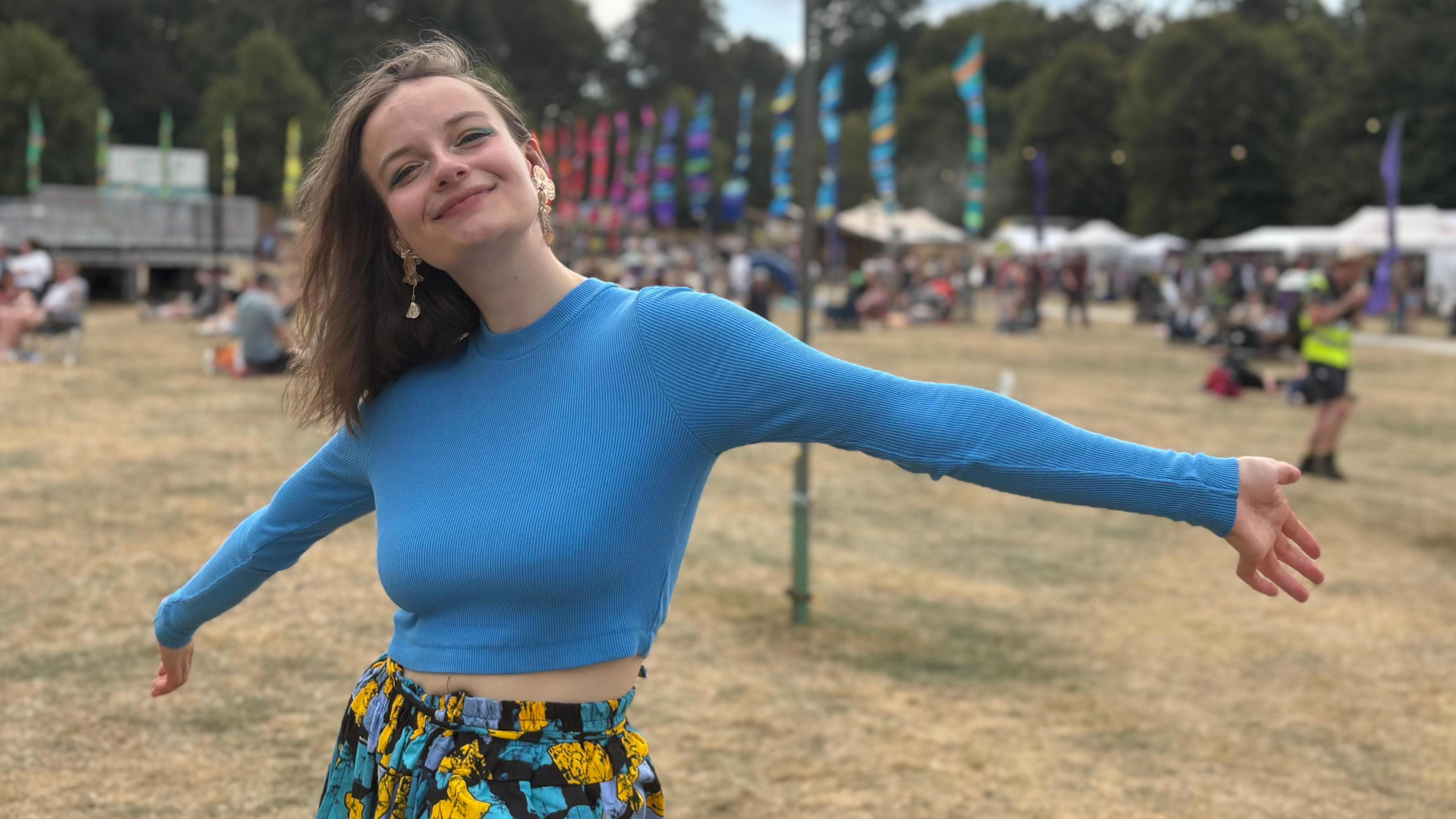 Sambambo, with long brown hair, wearing a blue long-sleeve top and blue and yellow skirt, smiling with her arms outstretched, standing on grass with festival-goers visible in the background.