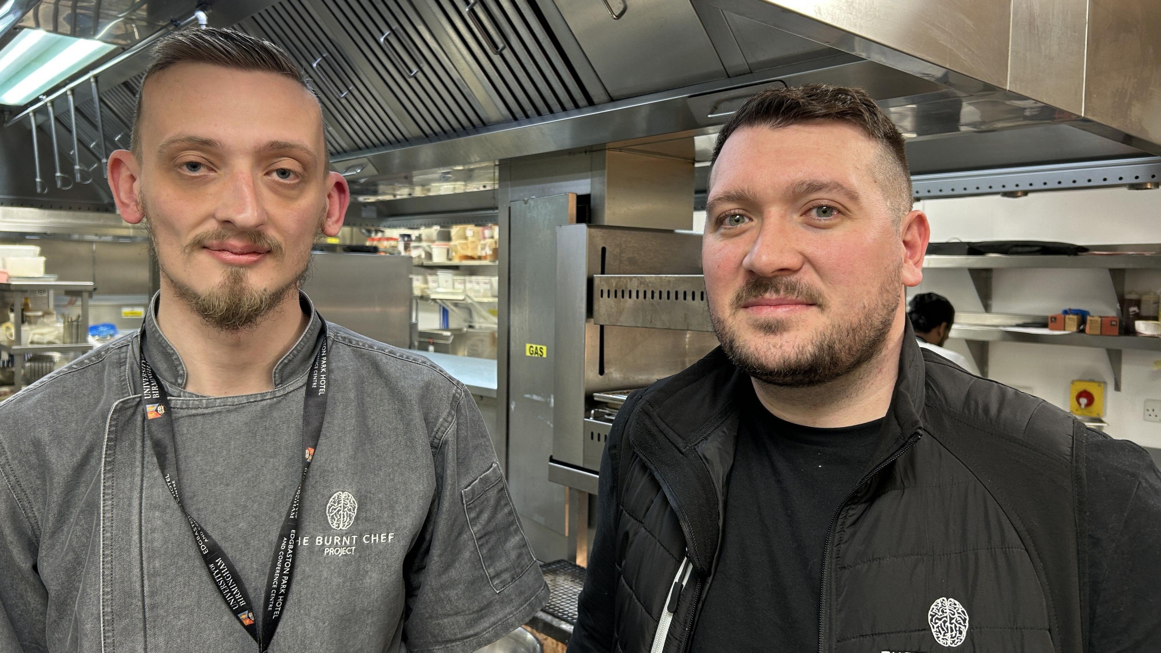 Two men are standing in a kitchen. On the left, Sam has short brown hair and brown facial hair. He is wearing a grey denim top that reads "the burnt chef project". Matt is standing next to him, he has short black hair and facial hair. He is wearing a black gilet with a logo on it